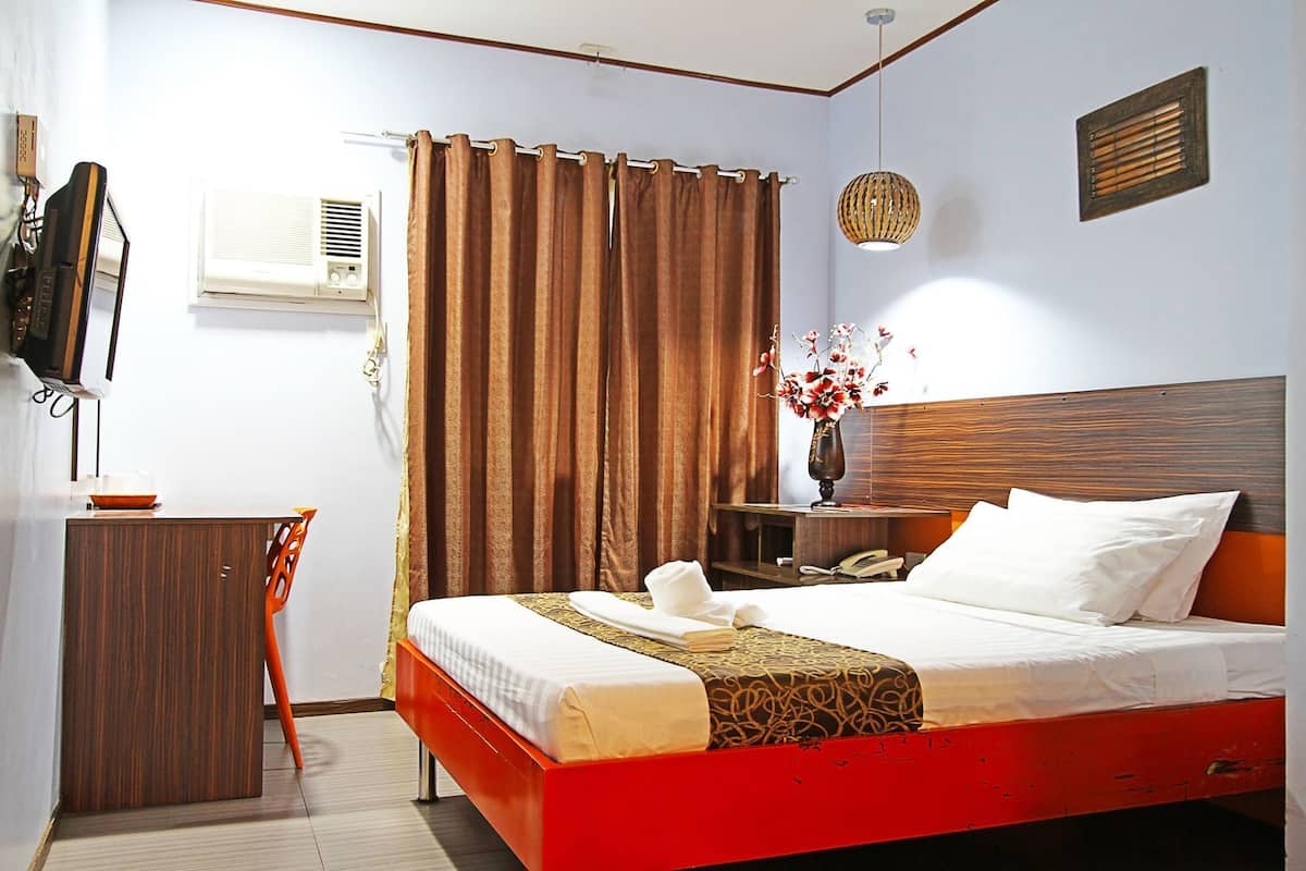 A hotel room with a bed, a desk, a chair, a TV, an air conditioning unit, a vase with flowers, and a wooden headboard.