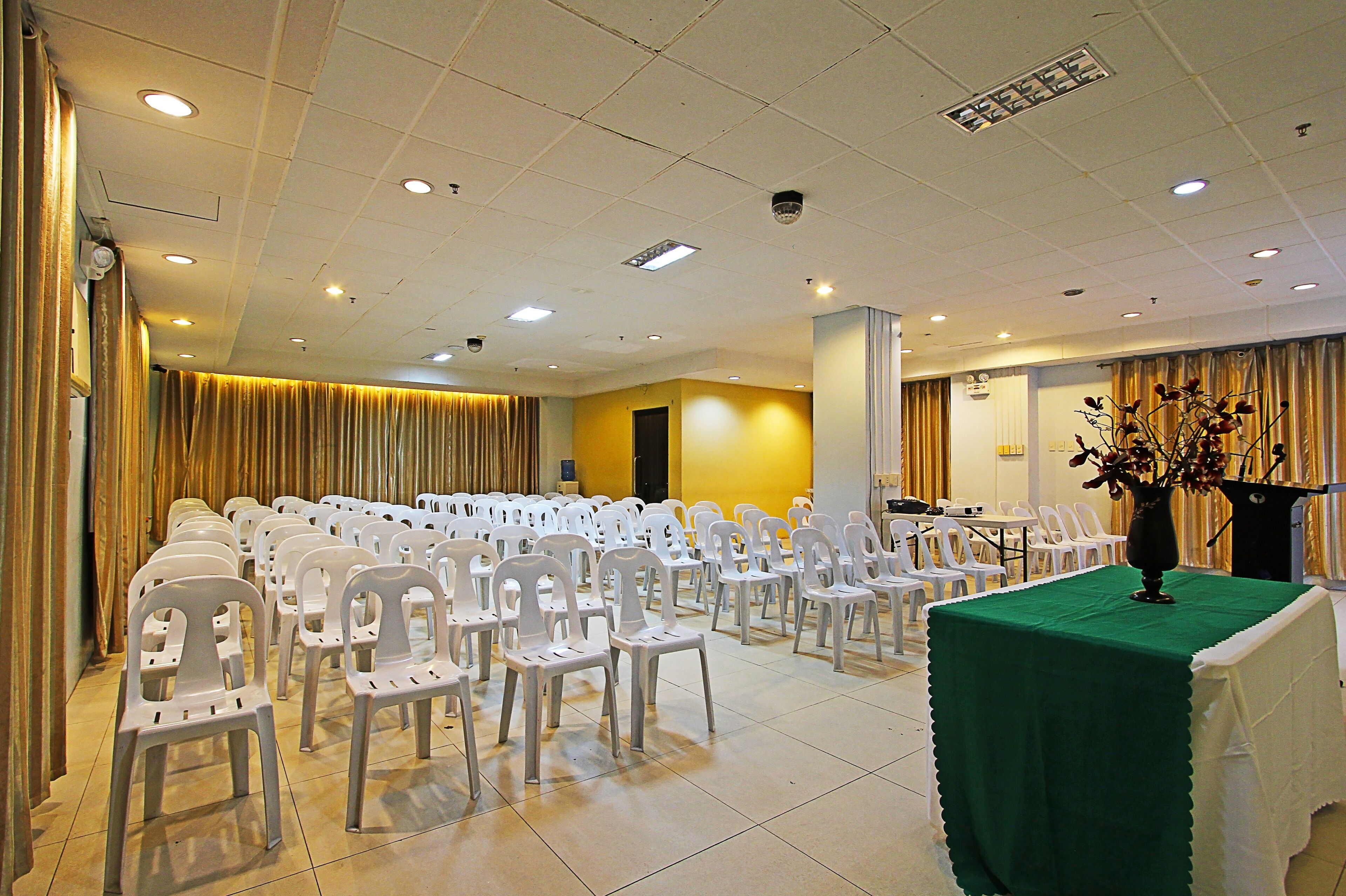 A conference room with rows of white plastic chairs, a green table with a vase, and a podium.