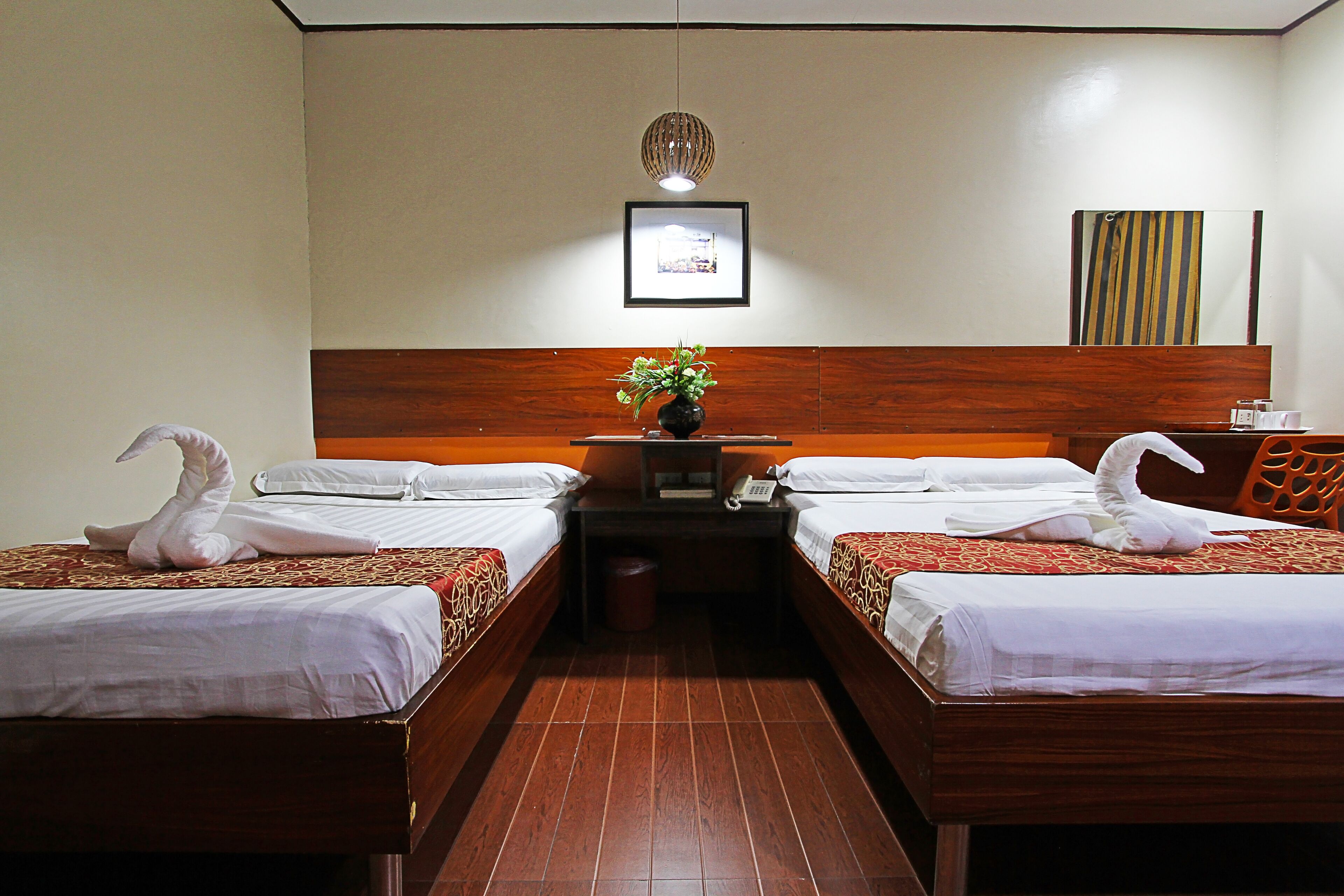 A hotel room with two beds, a wooden headboard, a small table with a vase of flowers, and a framed picture on the wall.