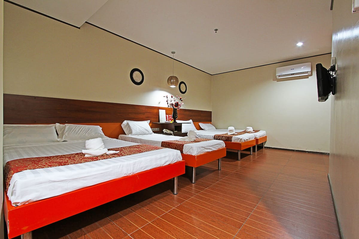 A hotel room with four beds arranged in a row, a wooden headboard, a nightstand with a lamp, and an air conditioning unit on the wall.