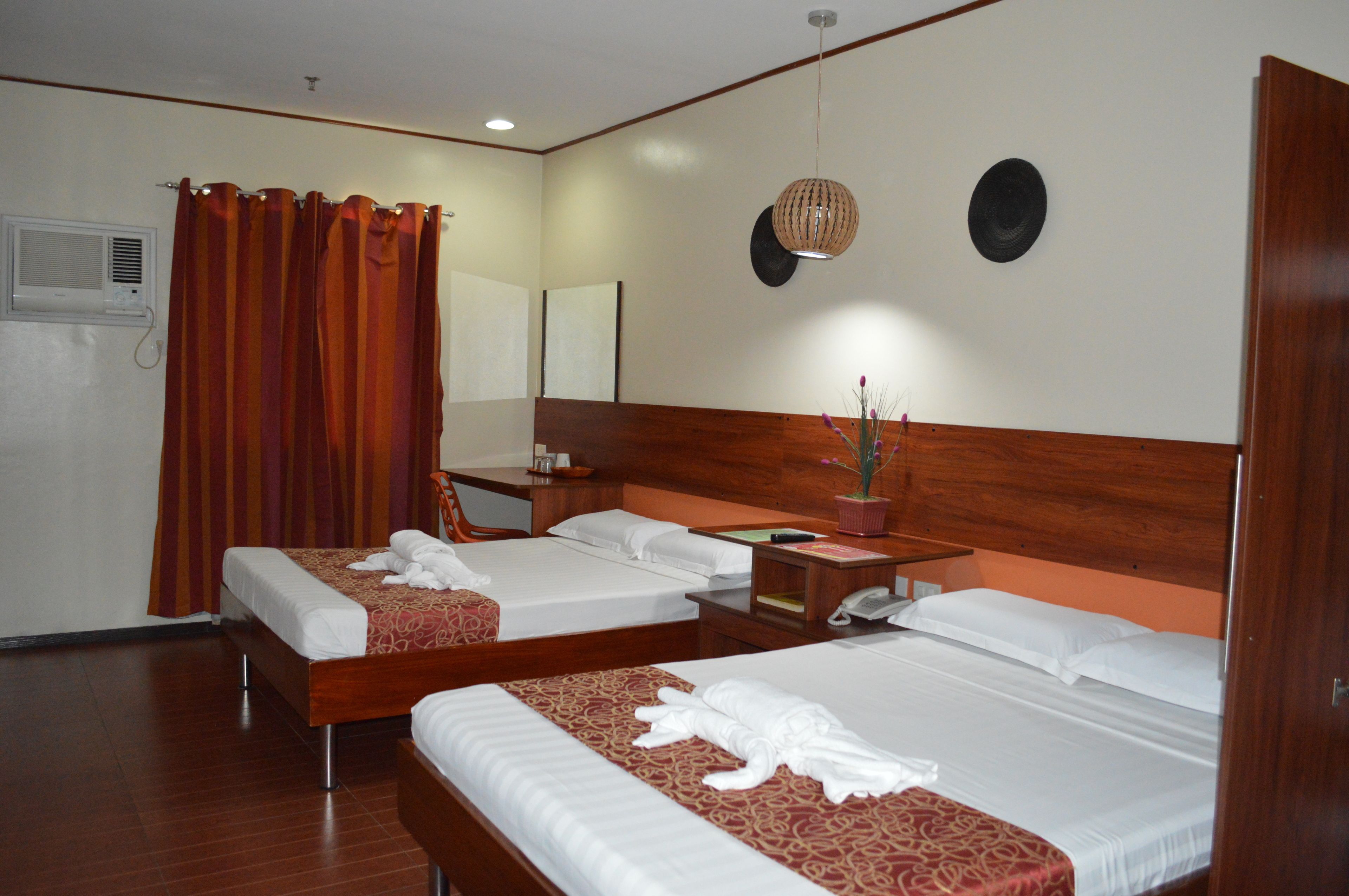 A hotel room with two beds, a wooden headboard, a bedside table with a lamp, a mirror, and a potted plant.