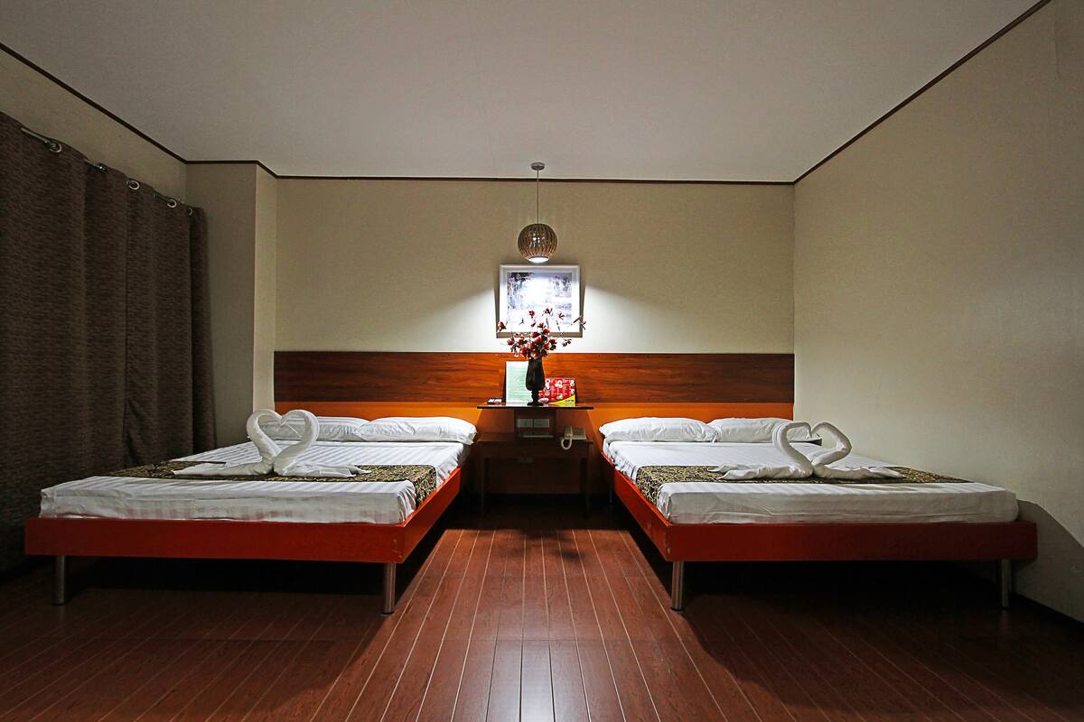 A hotel room with two single beds, a wooden headboard, a small table with a vase of flowers, and a ceiling light.