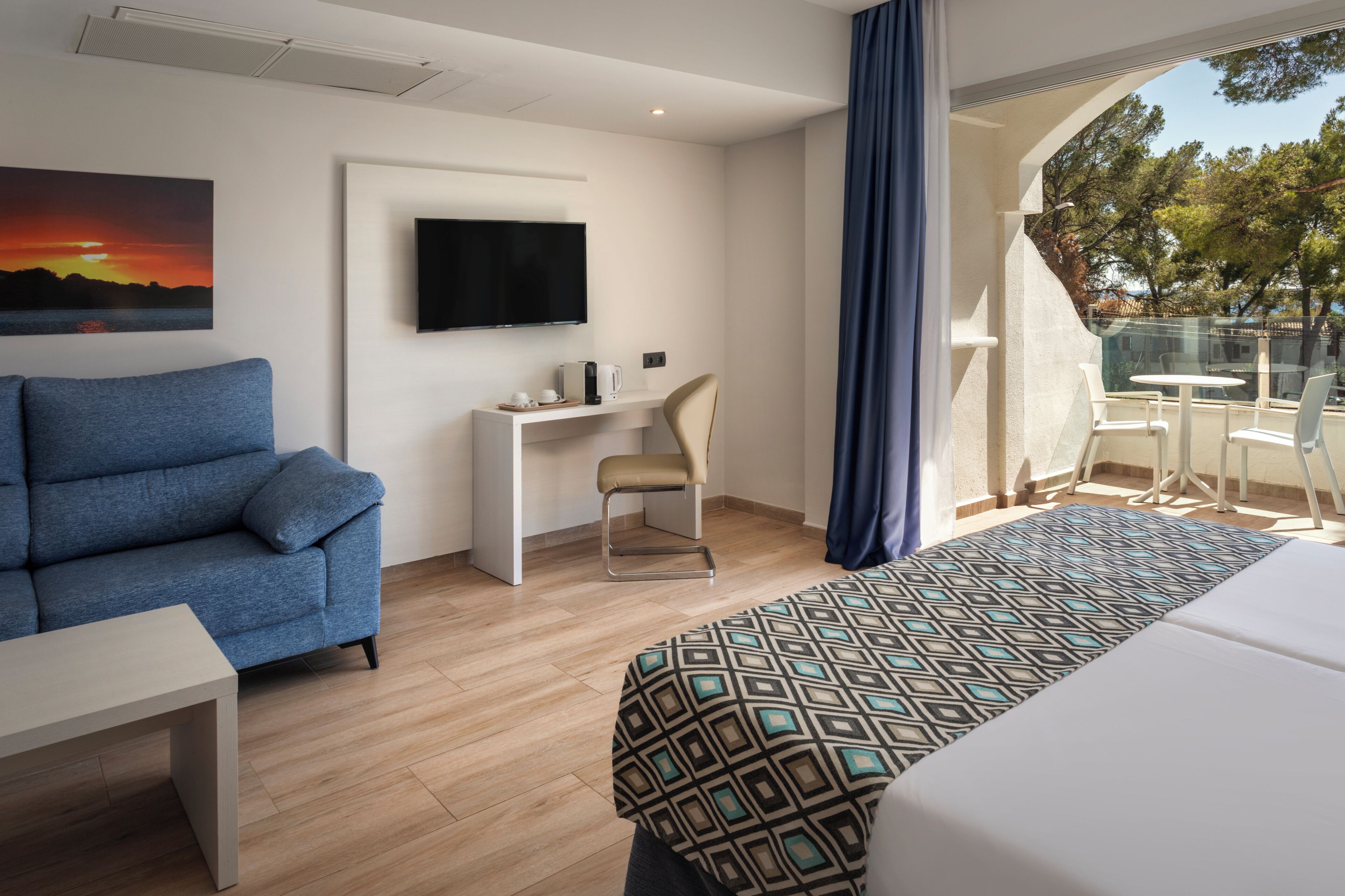A hotel room with a bed, a sofa, a desk, a chair, a TV, and a balcony with a view.