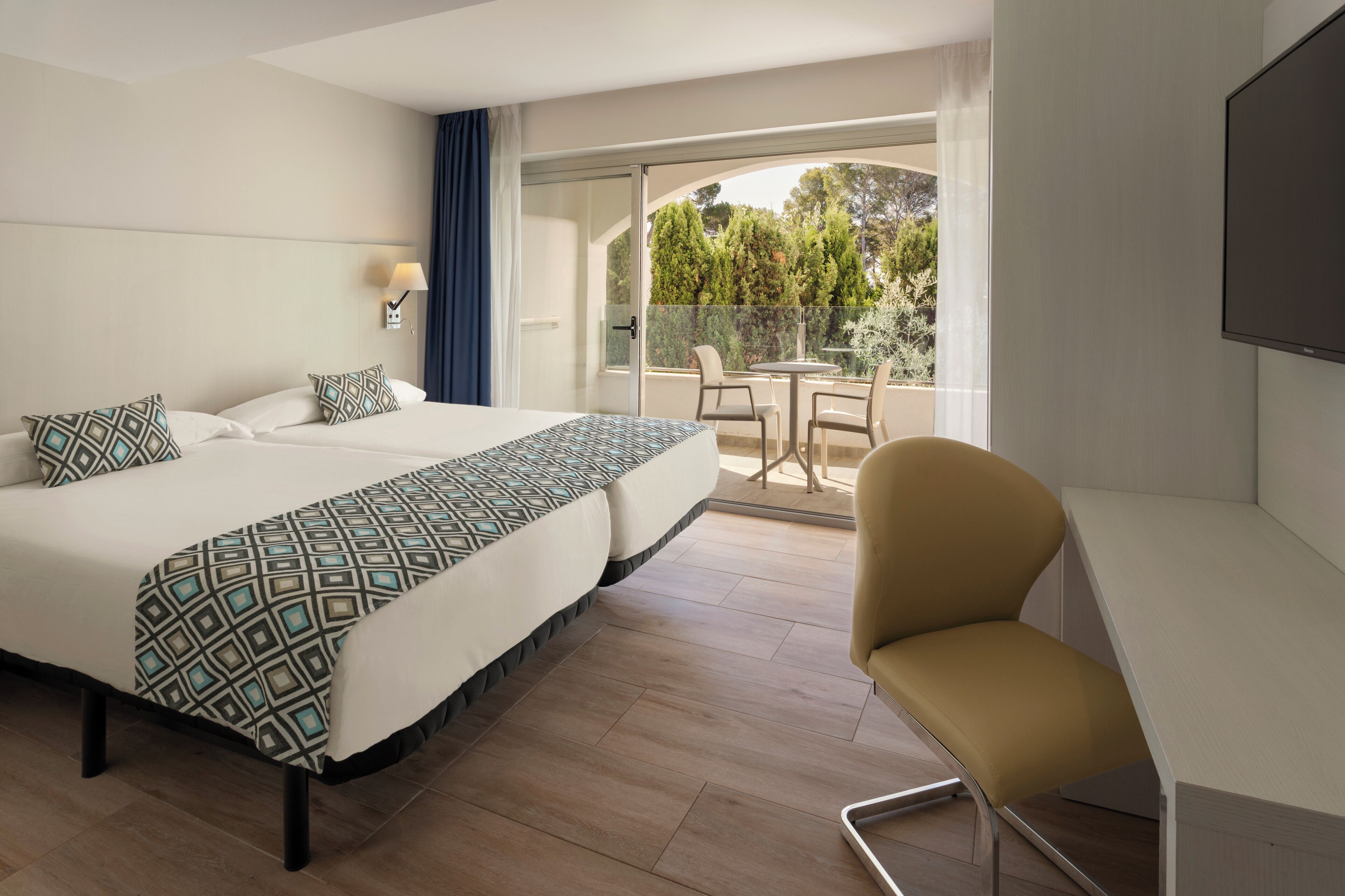 A hotel room with a bed, a desk, a chair, a TV, and a balcony with a table and chairs.