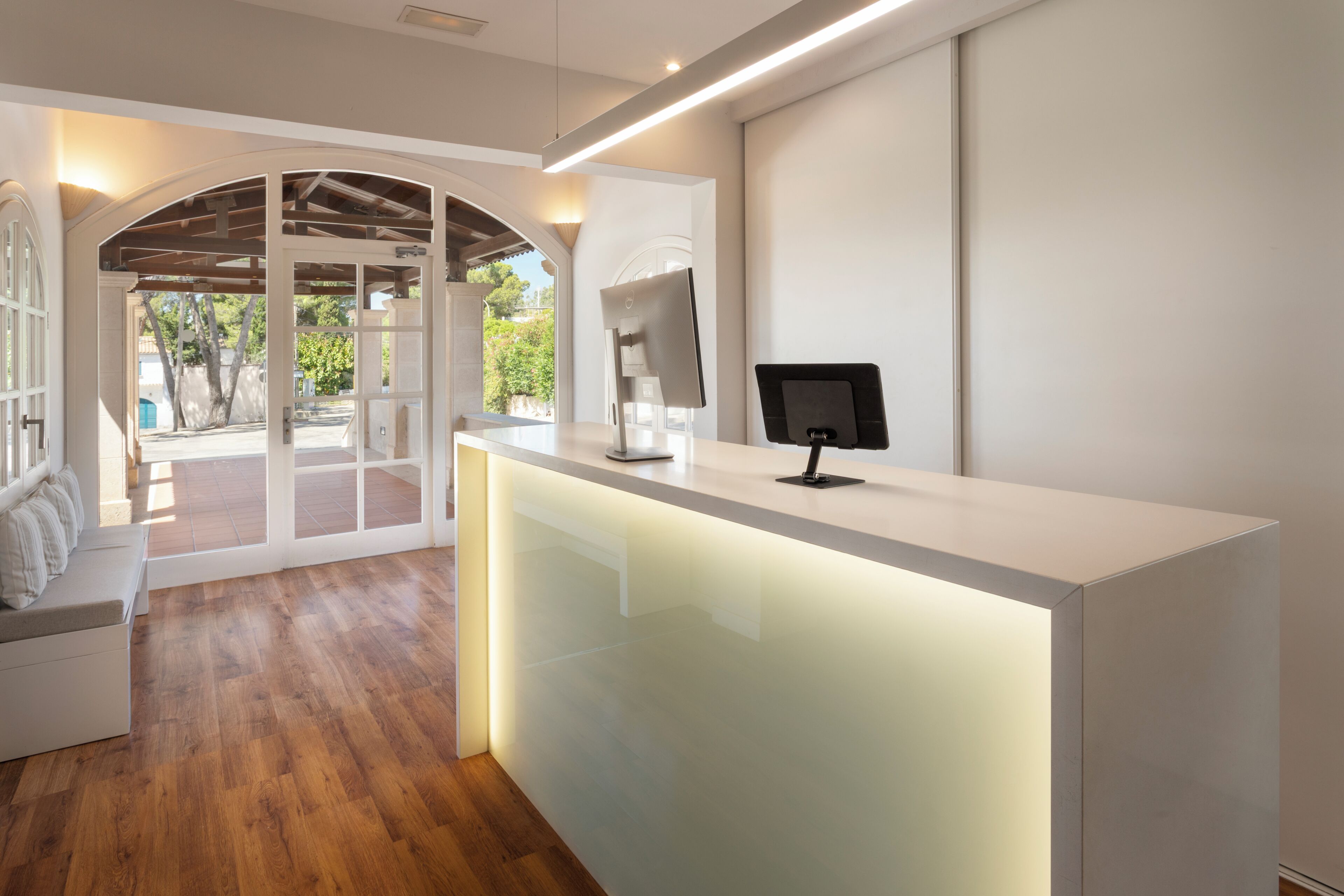 A reception desk with a computer monitor, a seating area with a bench, and a view of the outdoors through a glass door.
