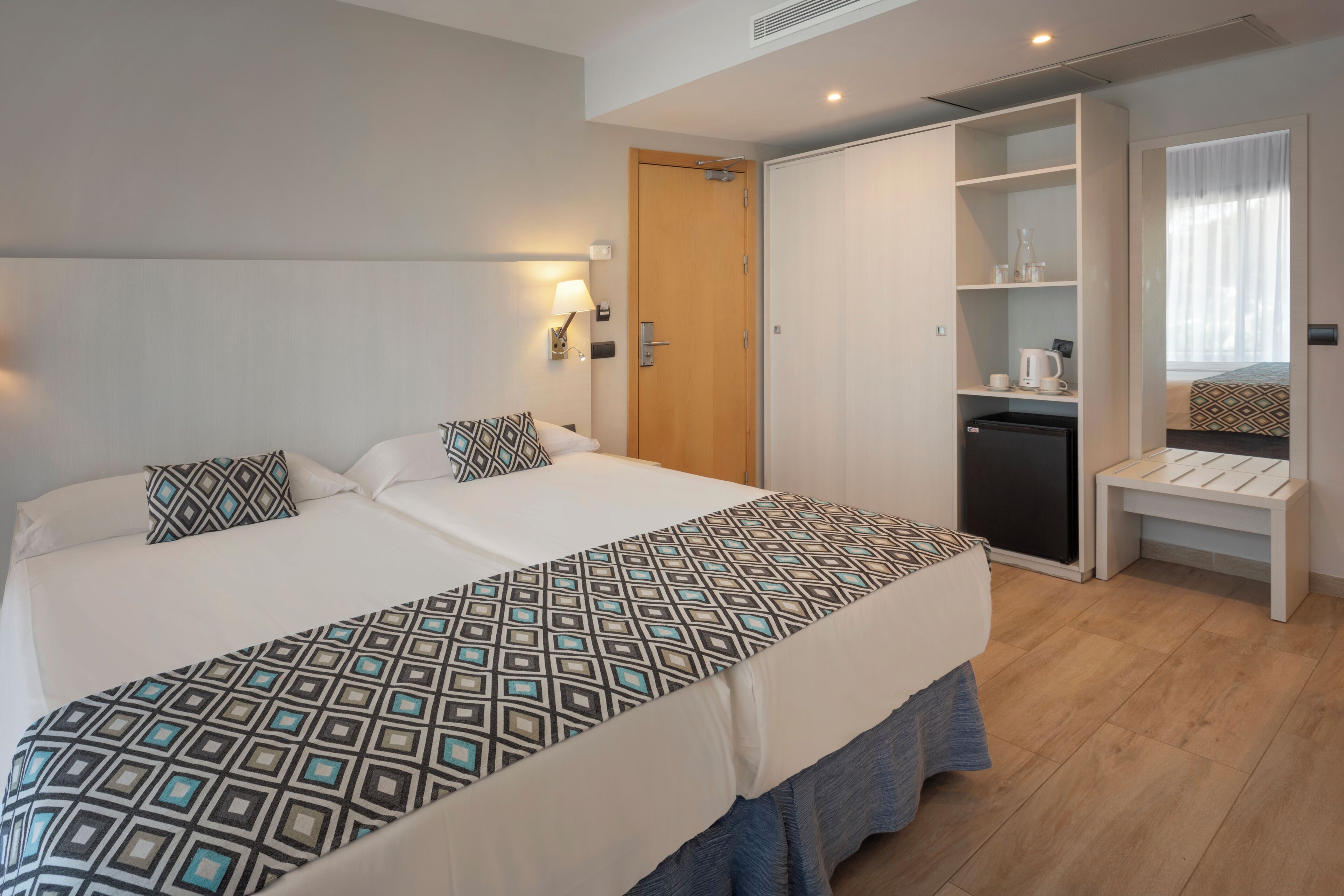 A double bed with patterned bedding, a bedside table, a wardrobe, a small kitchenette, and a mirror.