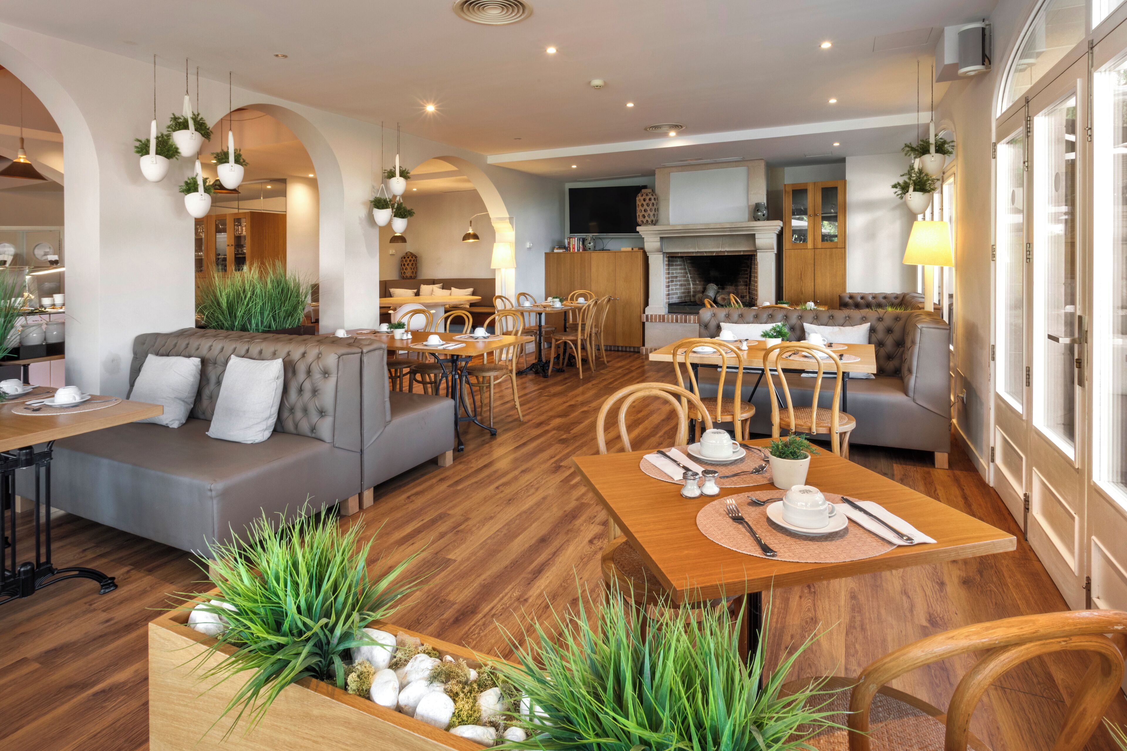 Un comedor moderno con mesas y sillas de madera, un sofá y plantas colgantes.