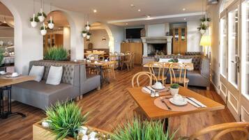 Un espace repas moderne avec des tables et des chaises en bois, un canapé et des plantes suspendues.