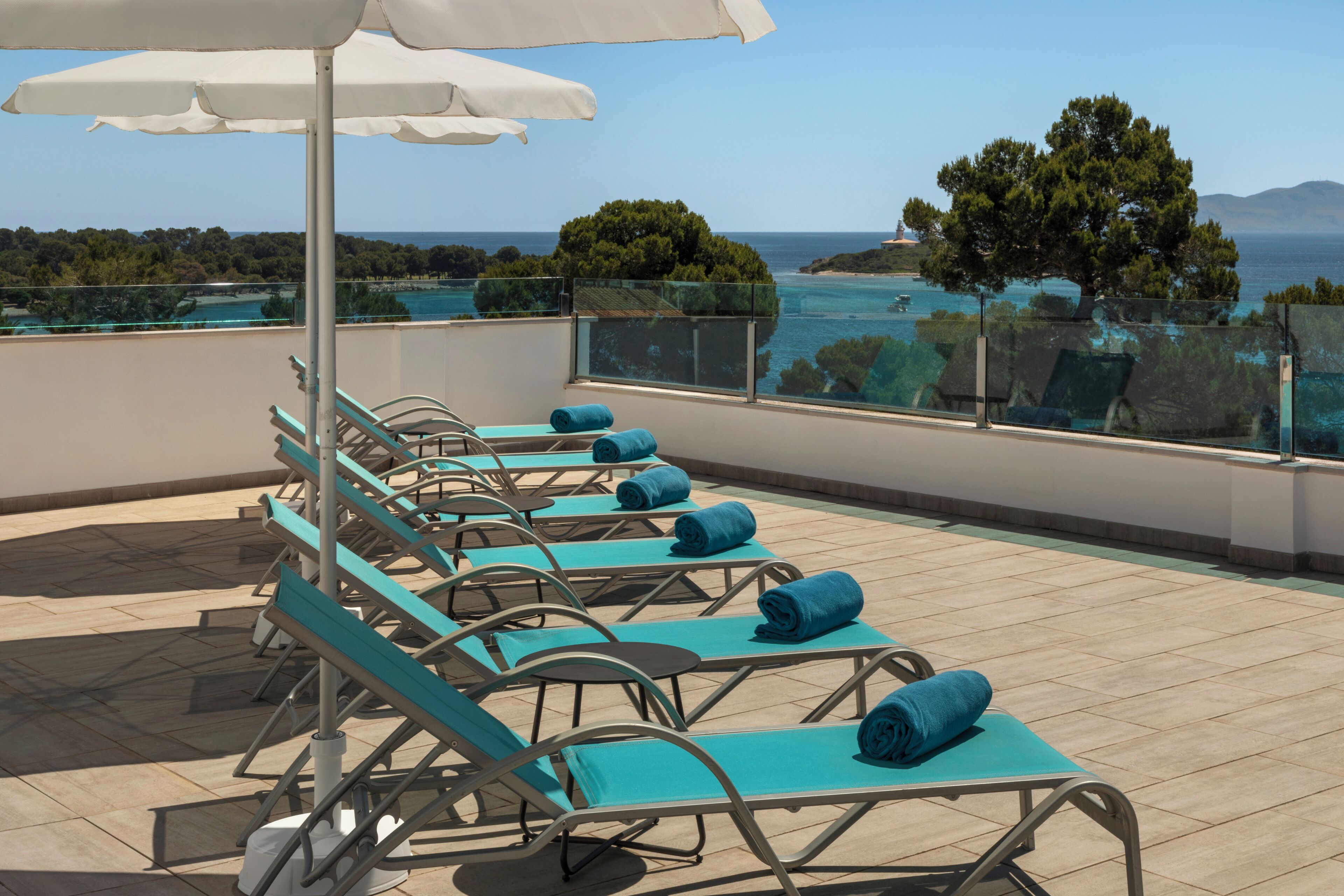A rooftop terrace with sun loungers, a white umbrella, and a view of the sea and trees.