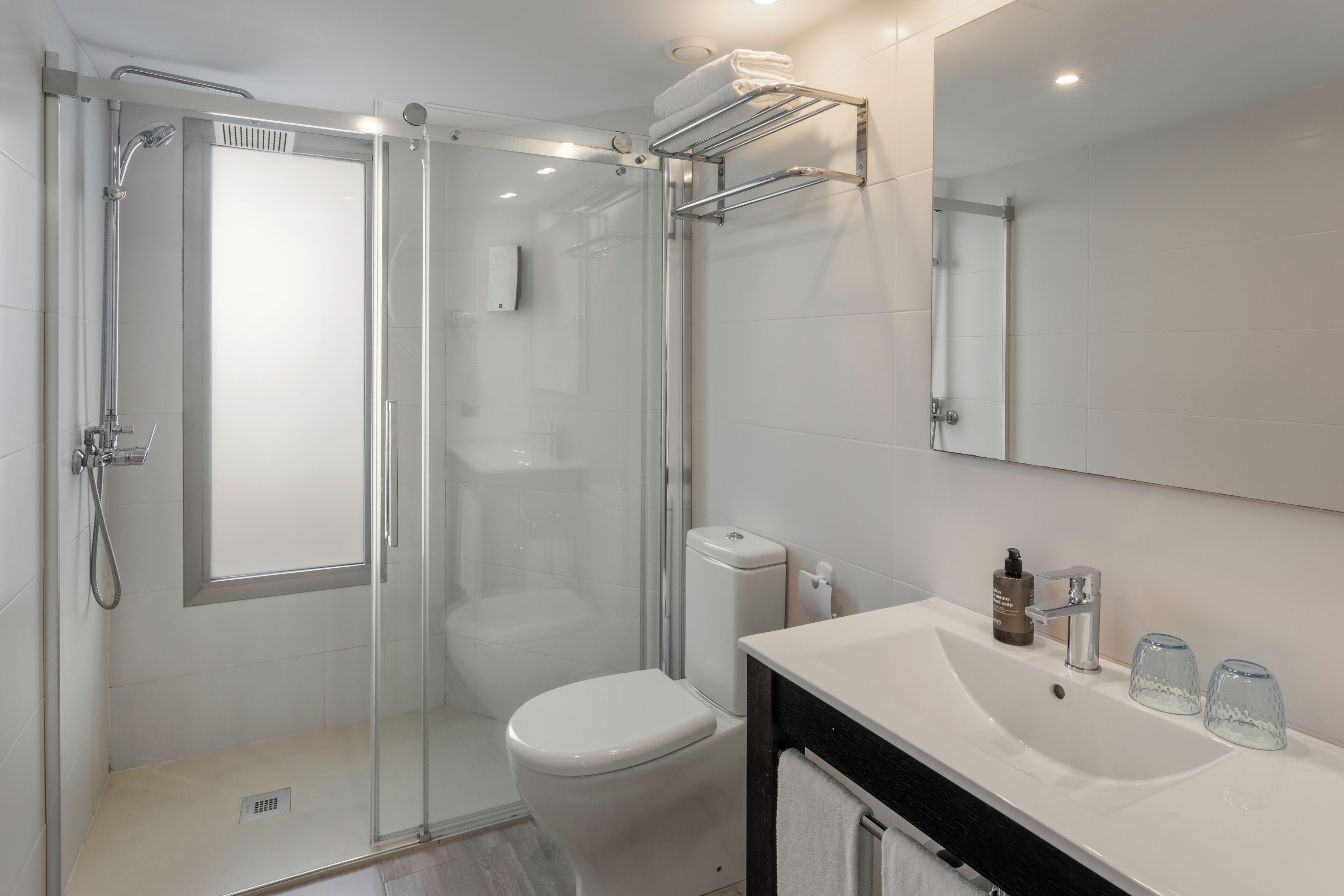 Un baño moderno con ducha de vidrio, inodoro blanco, lavamanos con espejo y un perchero para toallas.