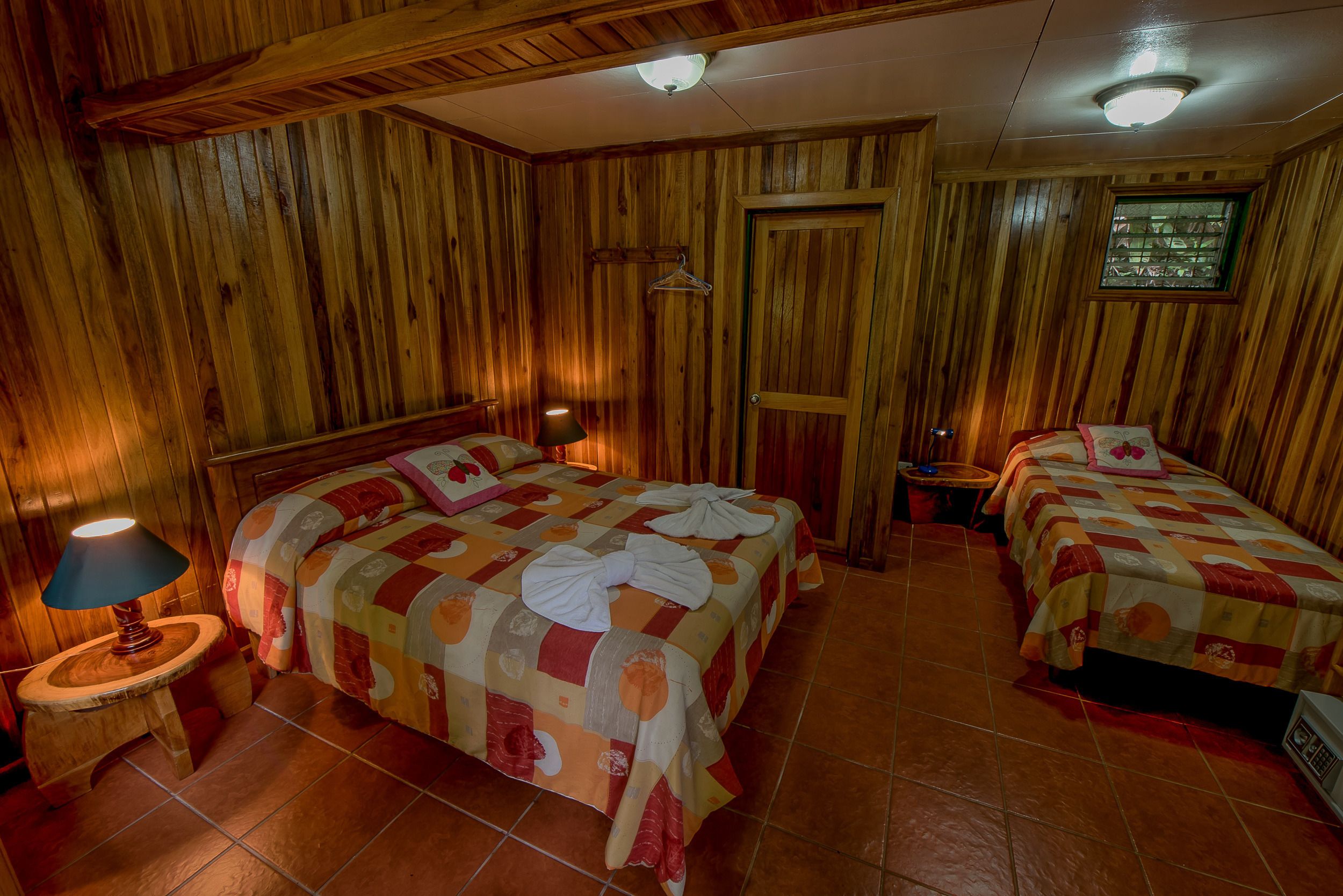 A wooden-paneled room with two beds, a hanging coat rack, and a small table with a lamp.