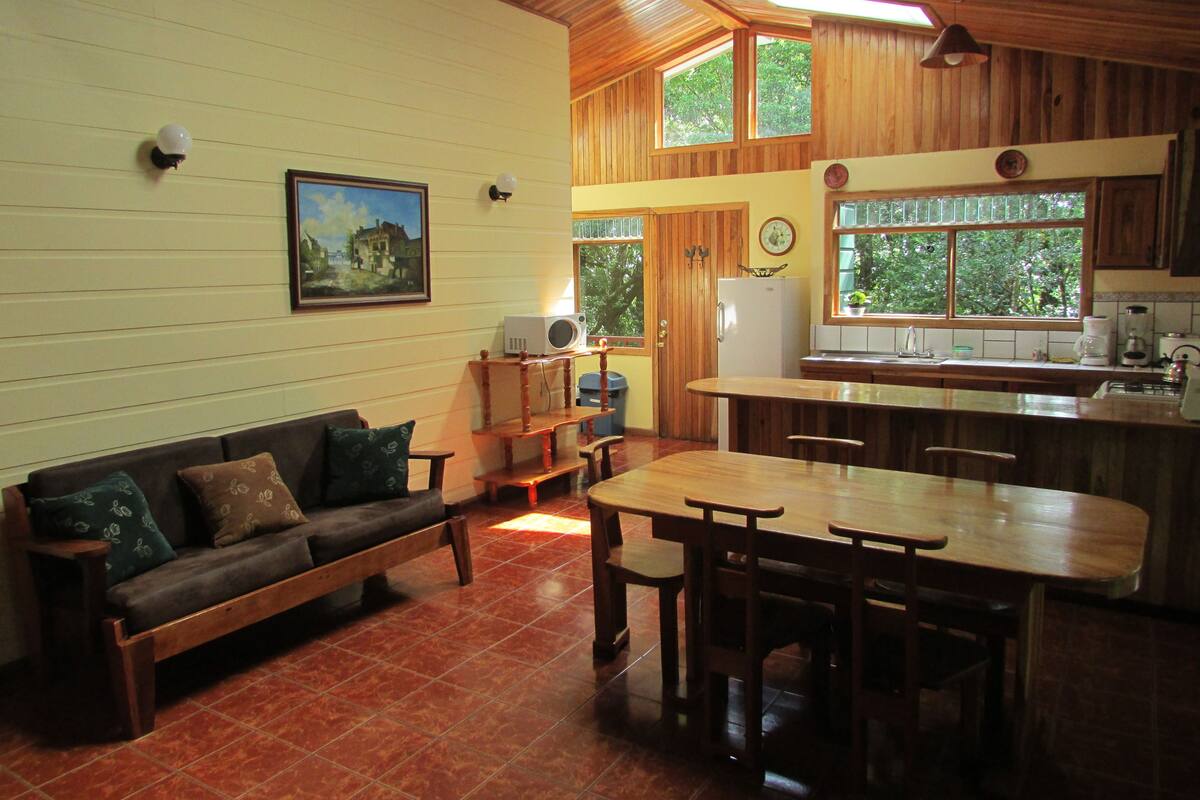 A cozy living room with a wooden bench, a dining table, and a kitchen area with a refrigerator and sink.