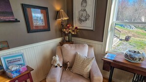A cozy living room with a cushioned armchair, a small table with a lamp, framed pictures, and a view of the outdoors.