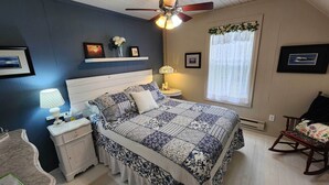 A bedroom with a bed, bedside tables, a nightstand, a lamp, a ceiling fan, a window with curtains, and a rocking chair.