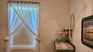 A room with a window covered by sheer curtains, a small table with a vase of flowers, and a framed picture on the wall.