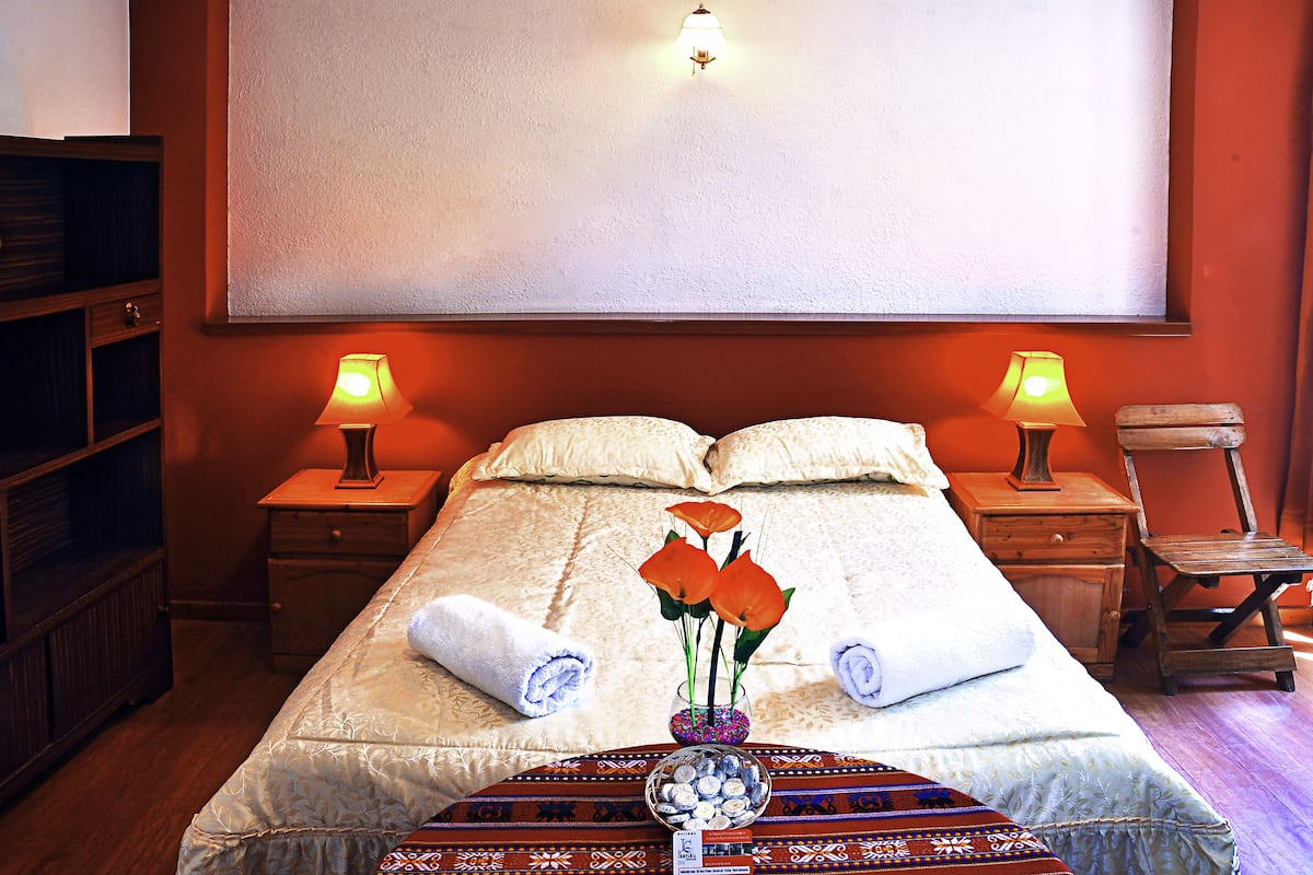Una cama bien tendida con dos almohadas, una mesita de noche con una lámpara, un pequeño jarrón con flores y una silla de madera.