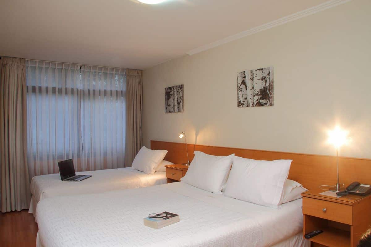 Ein Hotelzimmer mit zwei Betten, einem Laptop auf dem Bett links, einem Nachttisch mit Lampe und Telefon sowie einem Kunstwerk an der Wand.