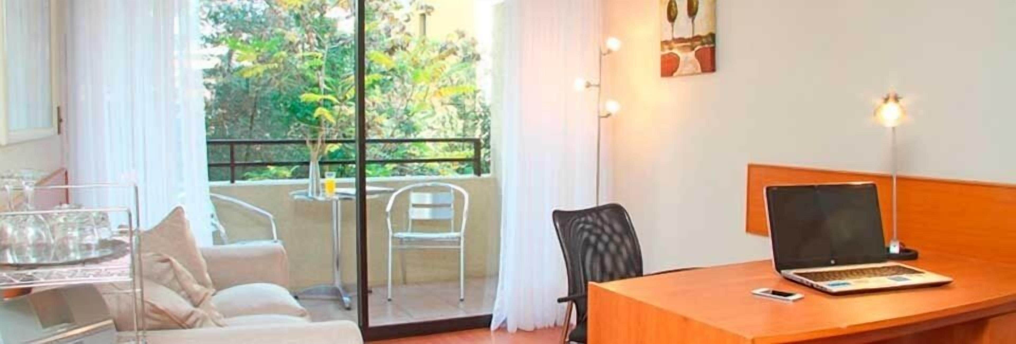 A hotel room with a balcony, a desk with a laptop, a chair, and a small table with a glass.