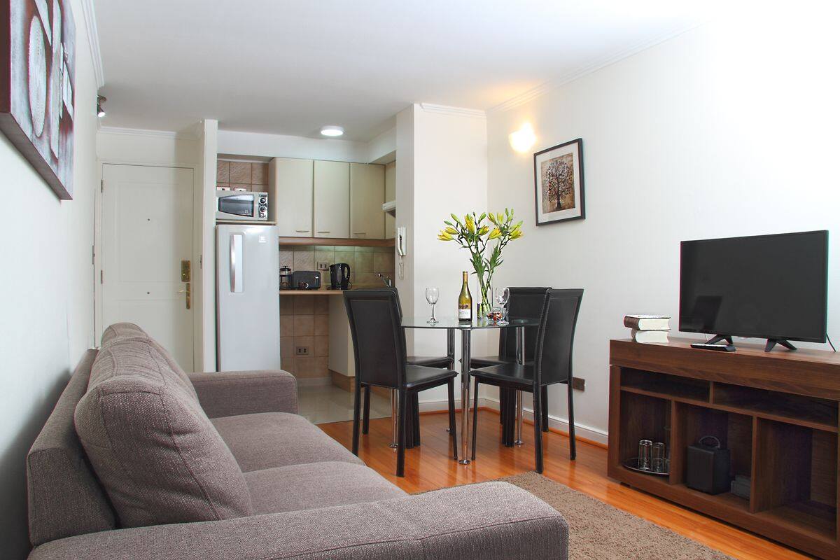Ein modernes Wohnzimmer mit einer Couch, einem Esstisch mit Stühlen, einem Fernseher auf einem Holzschrank und einer Küche im Hintergrund.