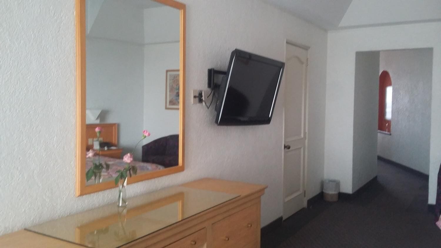 A room with a wooden dresser, a mirror, a television mounted on the wall, and a door leading to another room.