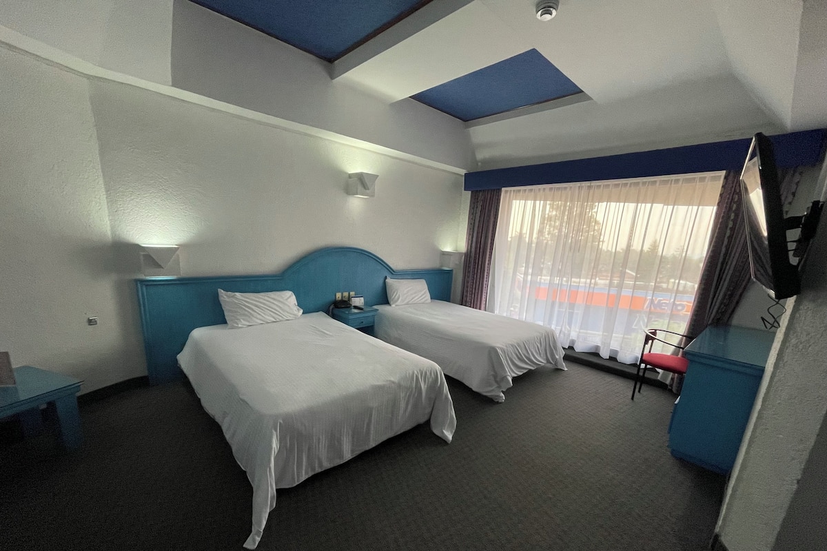 A hotel room with two single beds, a desk, a chair, and a large window with curtains.