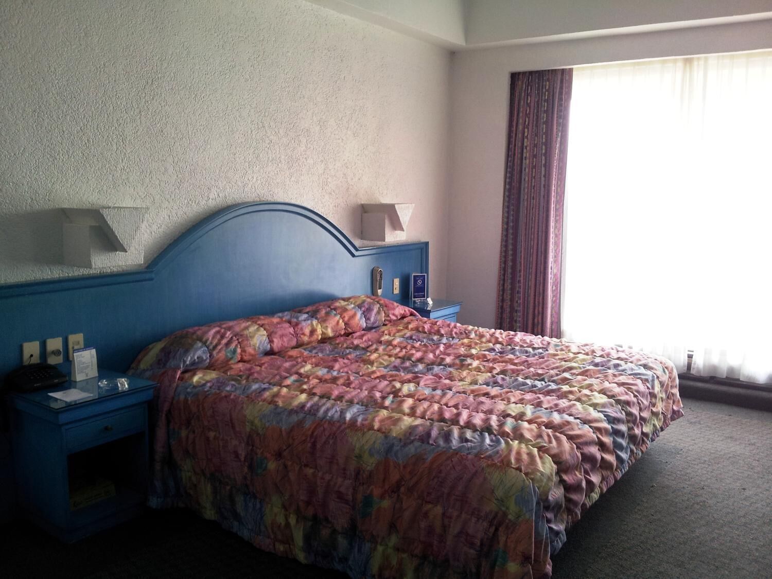 Habitación de hotel con una cama grande, una mesita de noche, una lámpara y un ventanal con cortinas.