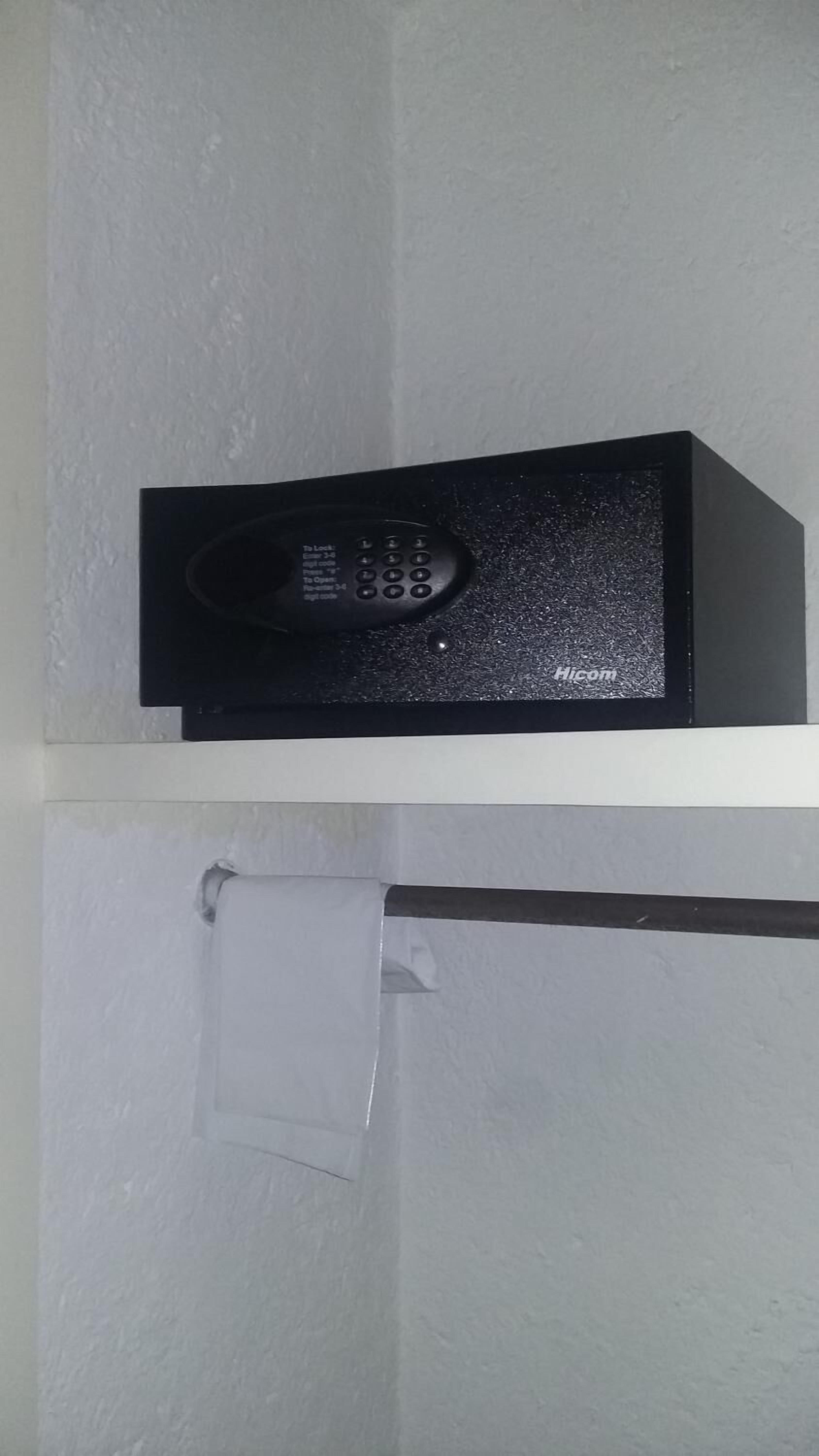 A black electronic device on a shelf, possibly a security system or intercom, with a white wall and a door with a towel hanging on it.
