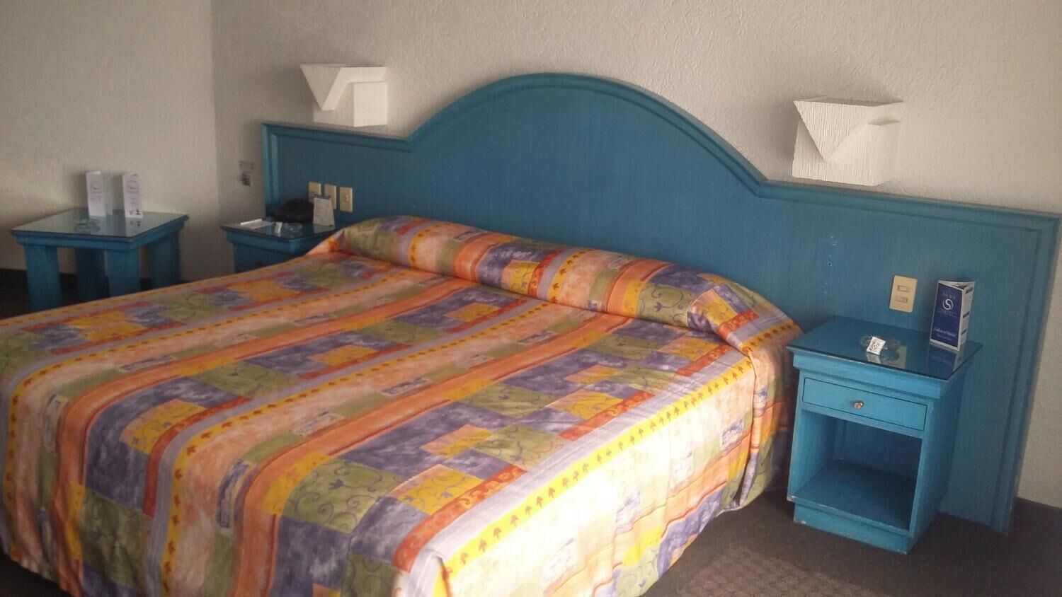 A bed with a colorful quilt, a blue headboard, and two bedside tables with lamps and tissues.