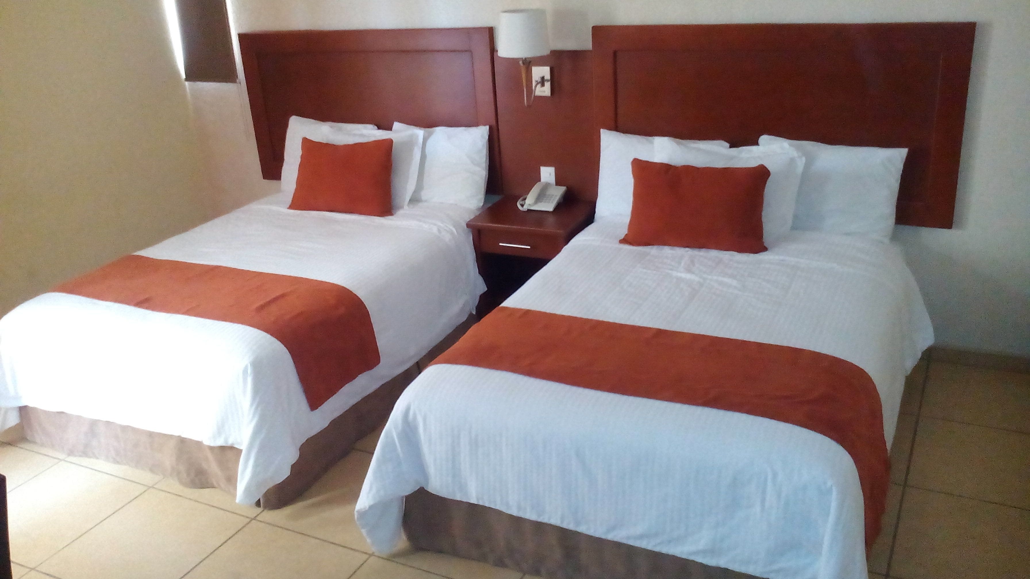 Duas camas de casal com roupa de cama branca e vermelha, uma mesa de cabeceira com telefone e uma luminária fixada na parede.