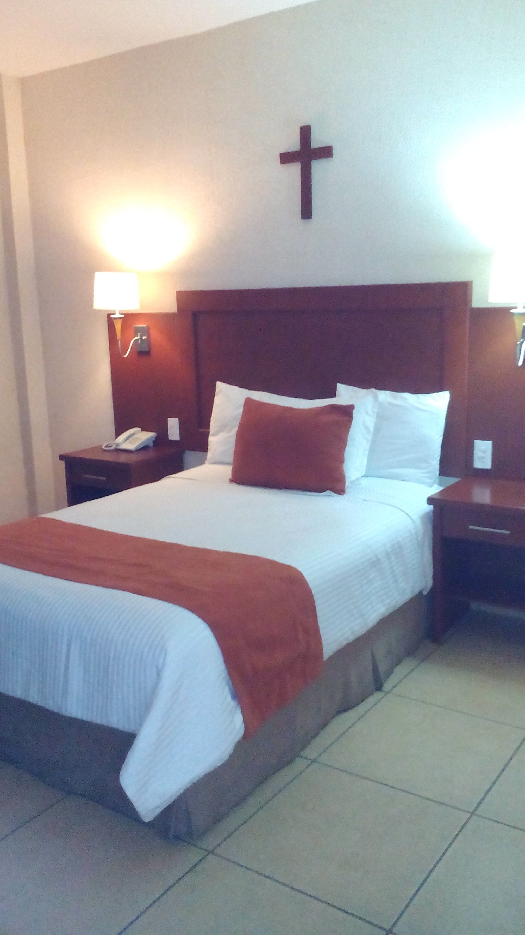Una habitación de hotel con una cama grande, una cruz en la pared y dos lámparas de noche.