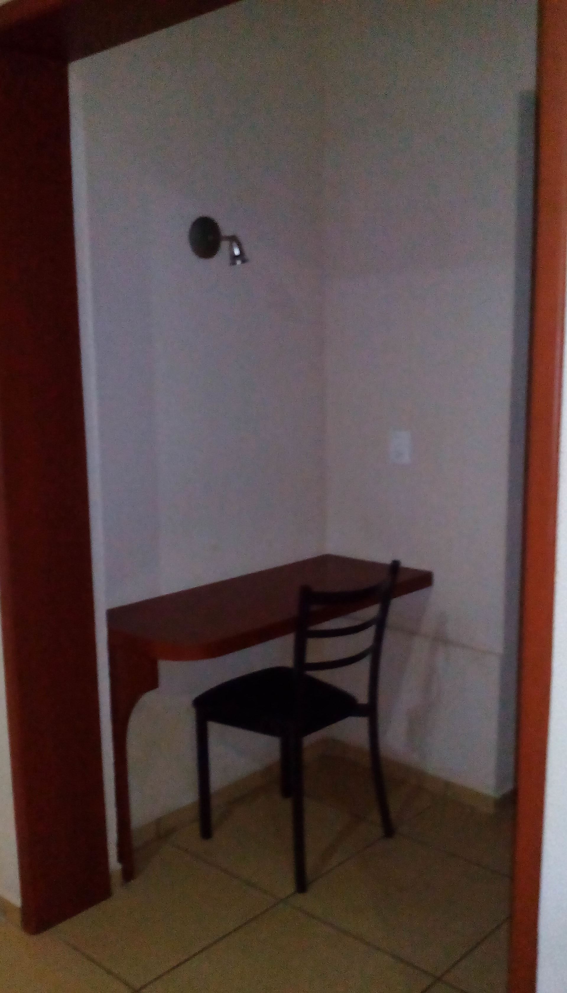 Una habitación pequeña, con poca luz, que tiene un escritorio y una silla de madera, un aplique en la pared y piso de baldosas.