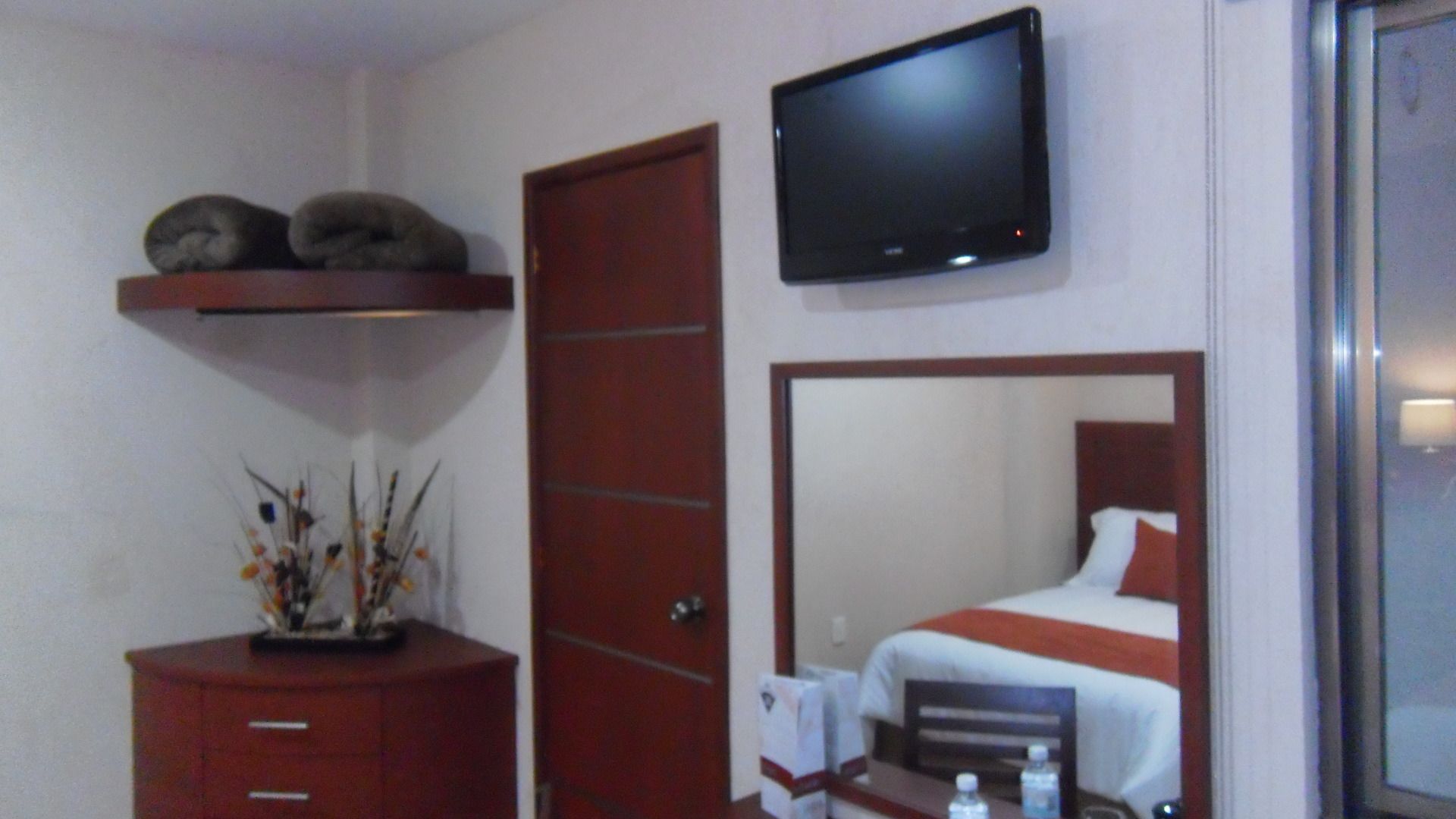 Habitación de hotel con una cama, un televisor montado en la pared, un aparador de madera y un espejo que refleja la cama.