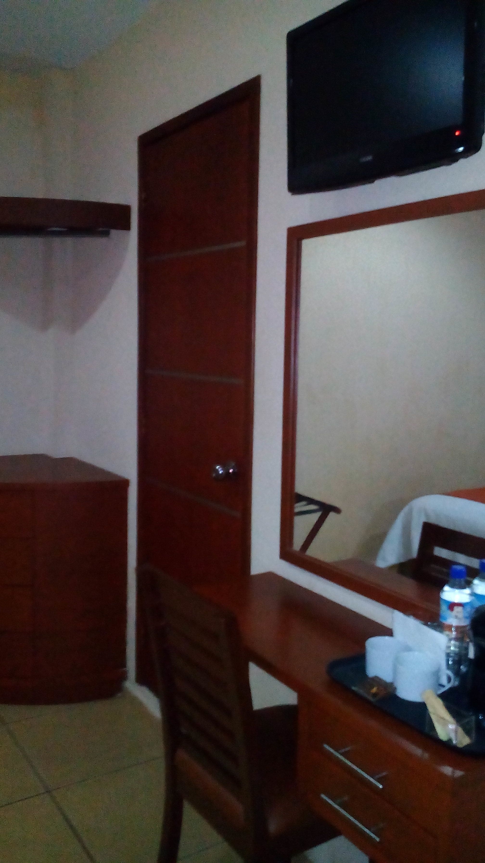 A room with a wooden desk, a chair, a mirror, a television mounted on the wall, and a closed door.