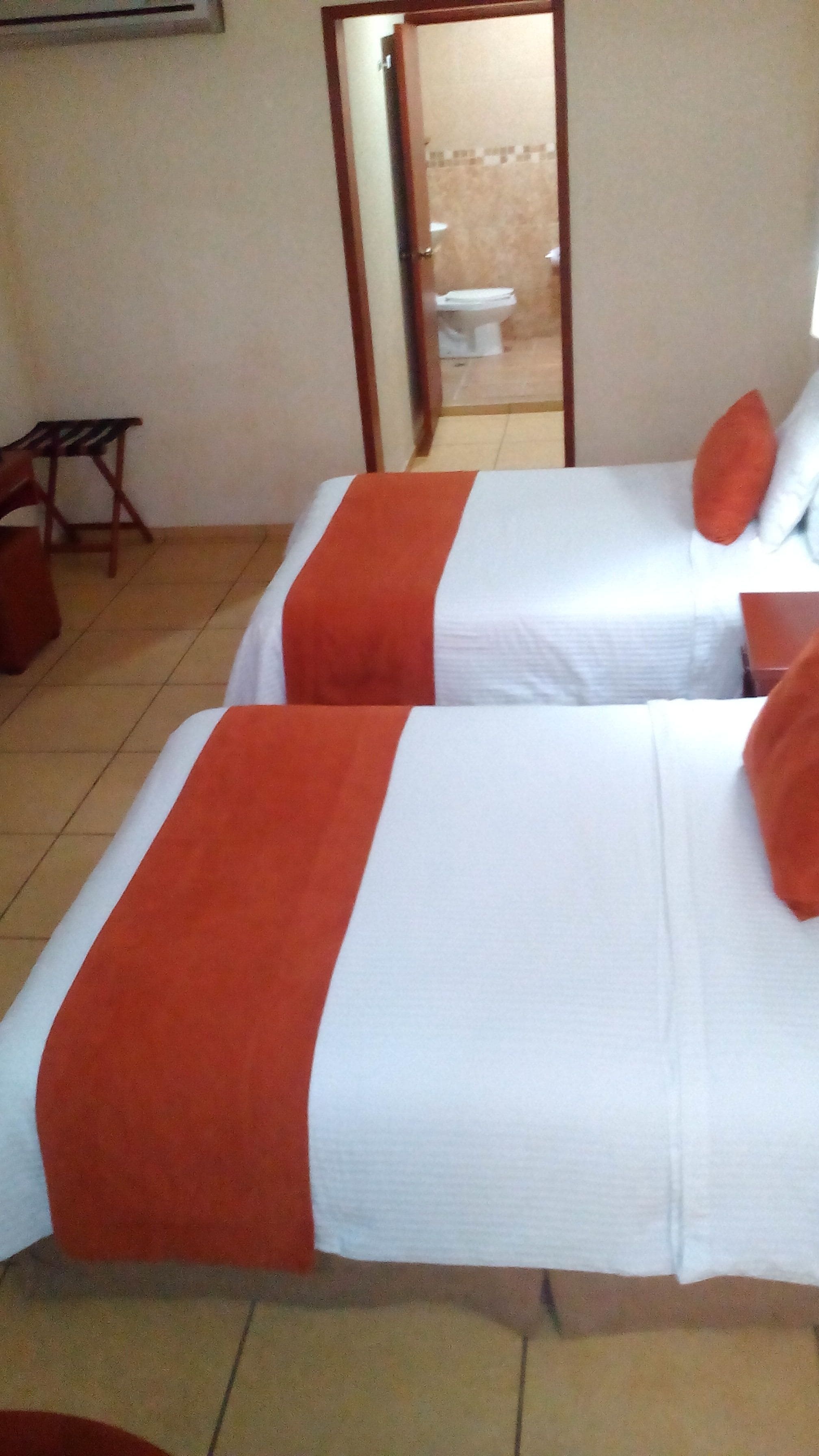 Dos camas con ropa de cama roja y blanca, una mesita de madera pequeña y un espejo que refleja un baño con un inodoro.