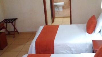Dos camas con ropa de cama roja y blanca, una mesita de madera pequeña y un espejo que refleja un baño con un inodoro.