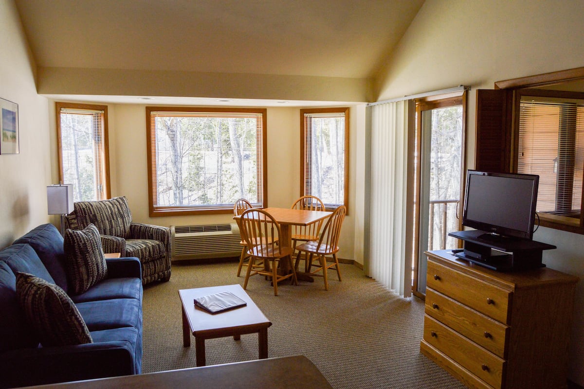 A room with a sofa, a dining table with chairs, a TV on a wooden cabinet, and large windows with blinds.