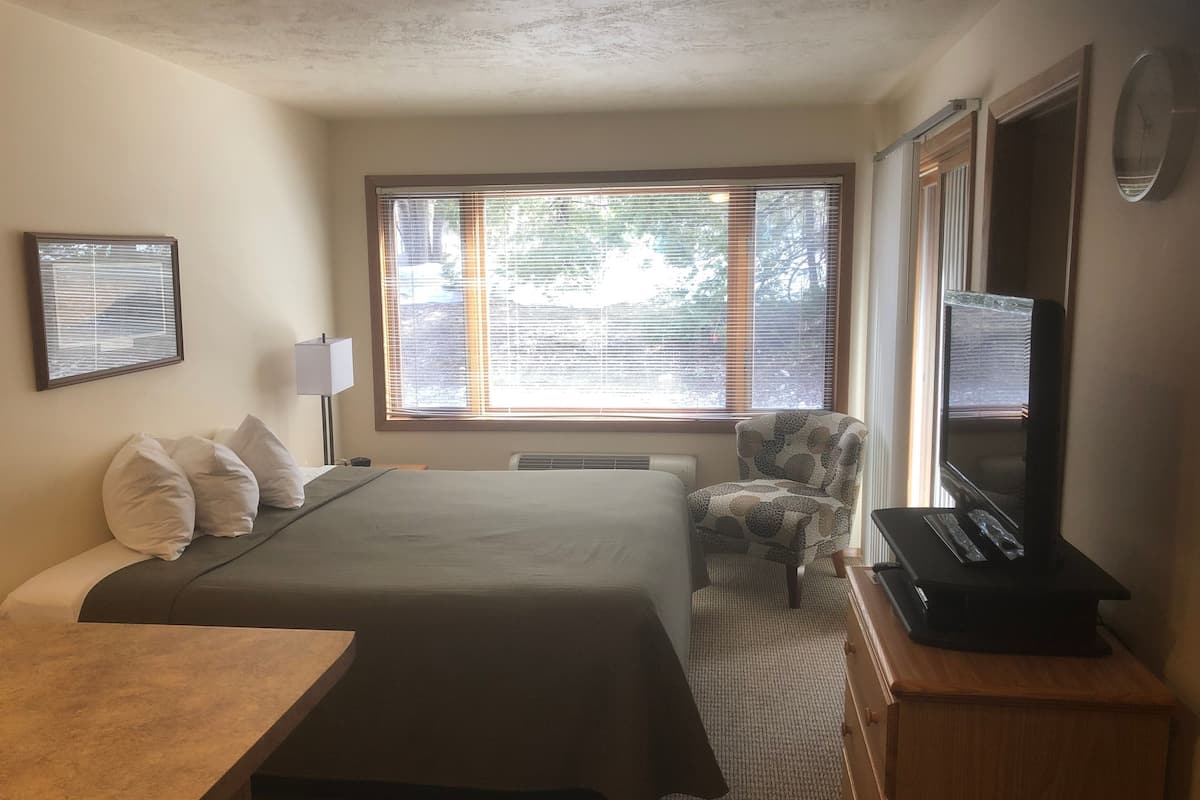 A hotel room with a bed, a chair, a desk, a TV, and a window with blinds.