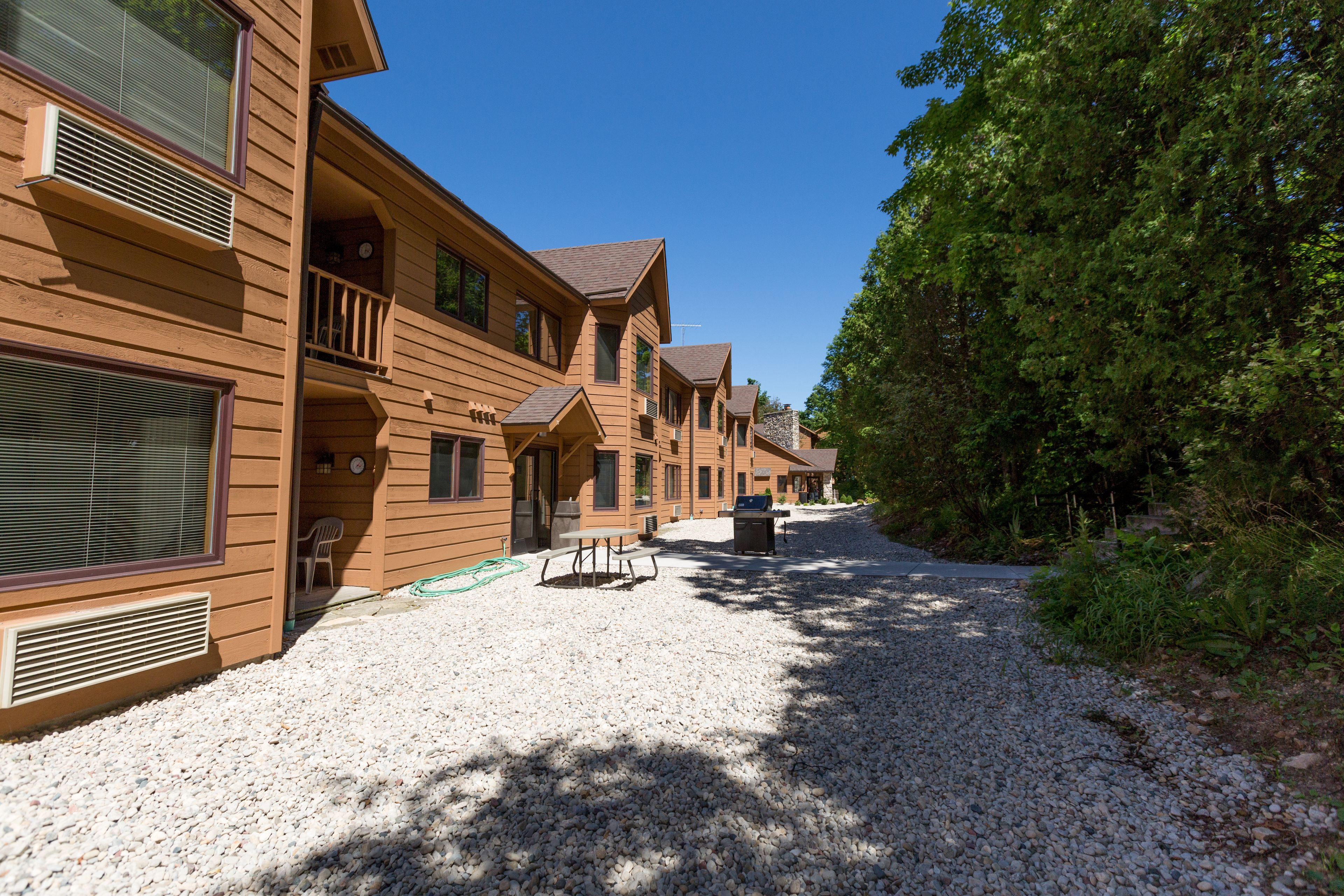 A row of wooden cabins with balconies, gravel driveway, and surrounding trees.