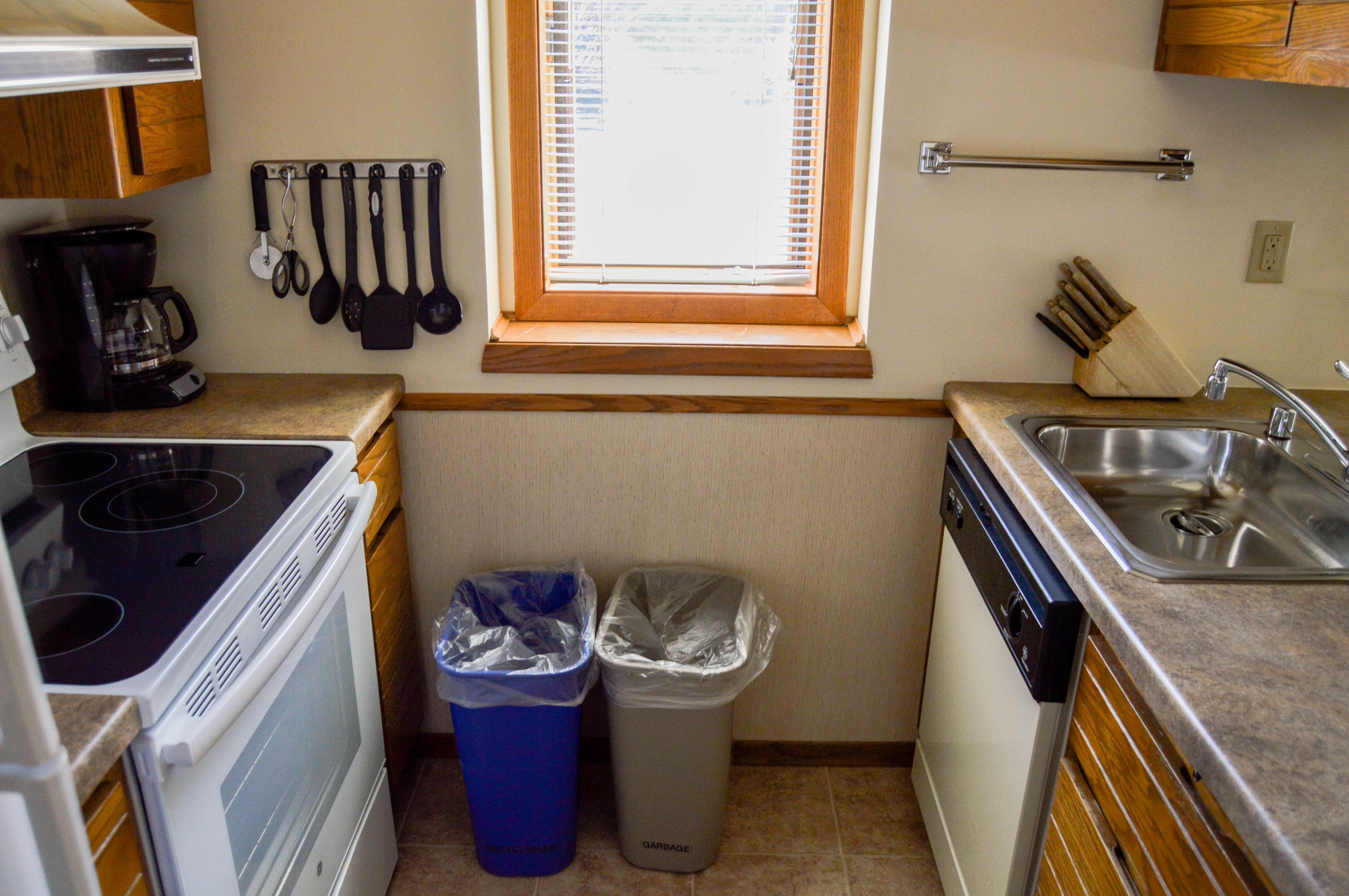 A compact kitchen with a stove, dishwasher, and sink. There are two trash bins, a window with blinds, and a knife block on the countertop.