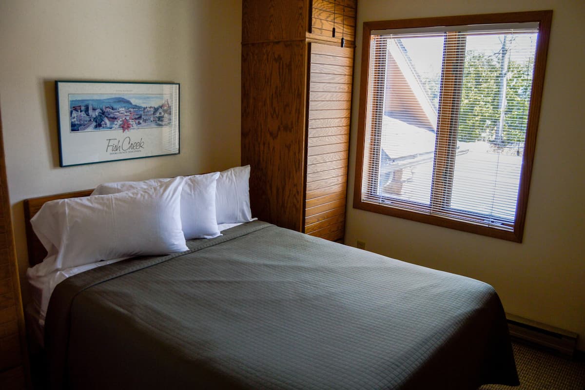 A neatly made bed with white pillows and a gray bedspread in a room with a wooden headboard, a framed picture of a town on the wall, and a window with blinds.
