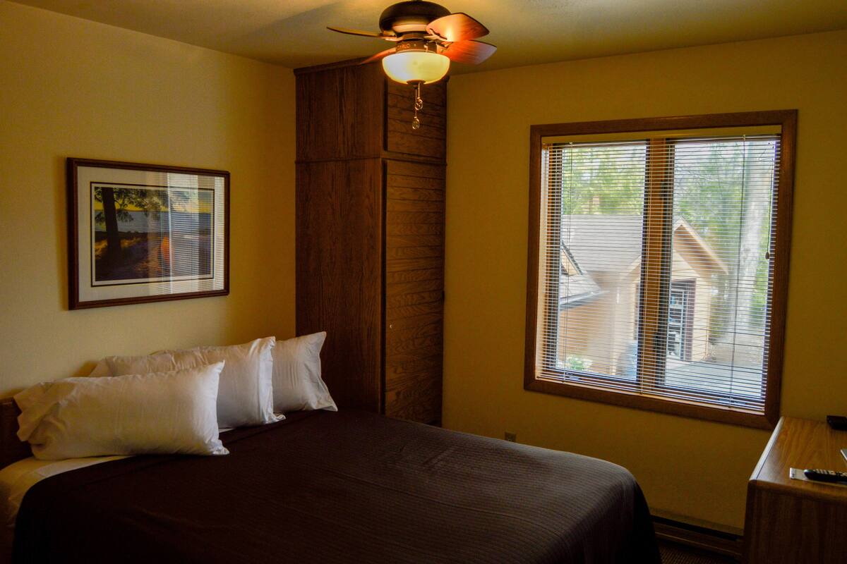 A hotel room with a bed, a ceiling fan, a window with blinds, and a framed picture on the wall.