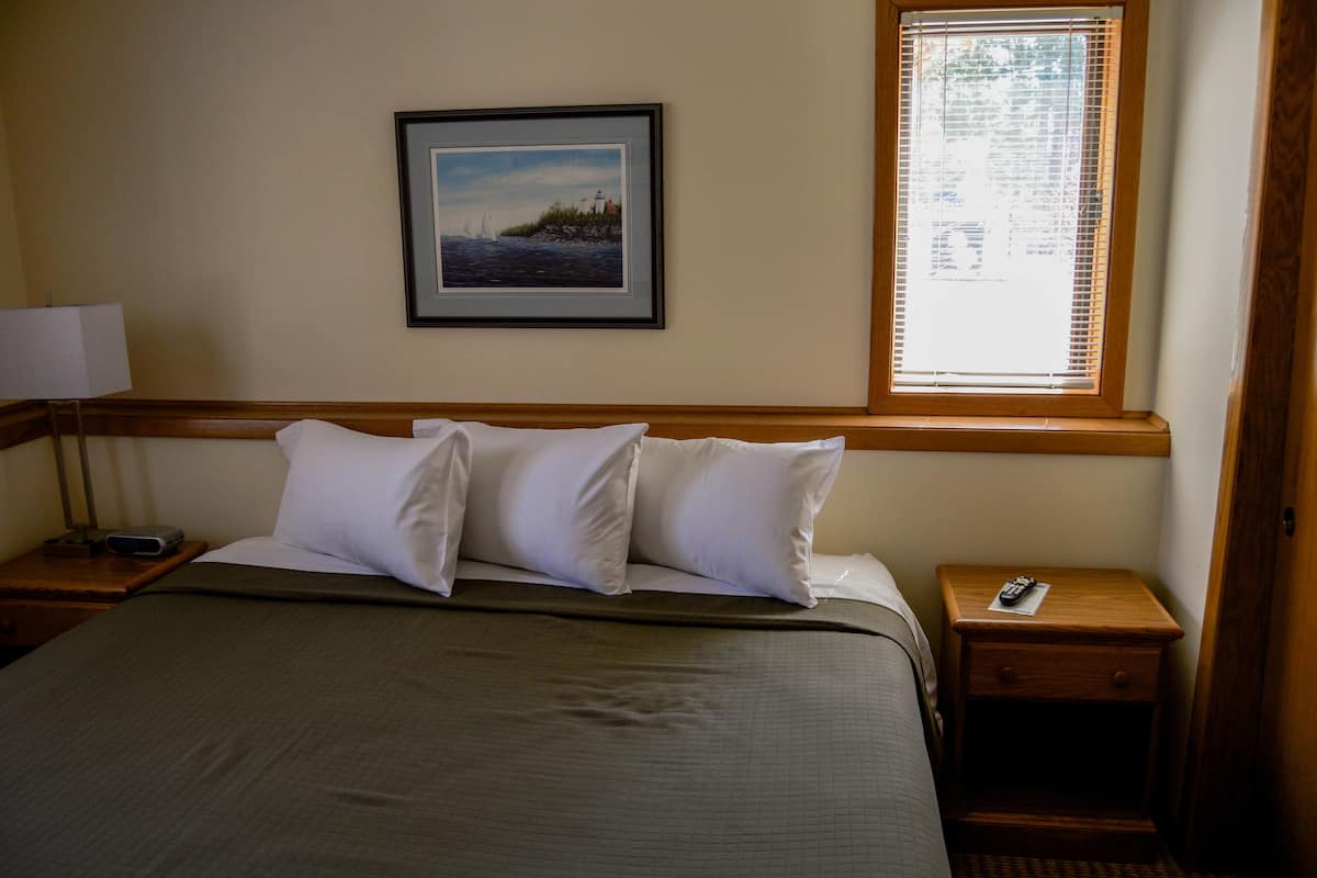 A neatly made bed with white pillows and a green bedspread in a hotel room. There is a framed picture on the wall above the bed, a window with blinds, and a nightstand with a lamp and remote control.
