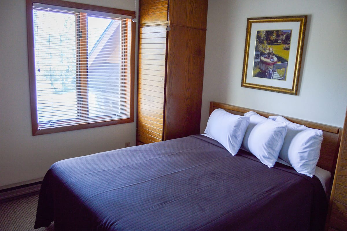 A neatly made bed with a dark-colored bedspread and white pillows in a room with a large window and a framed picture on the wall.