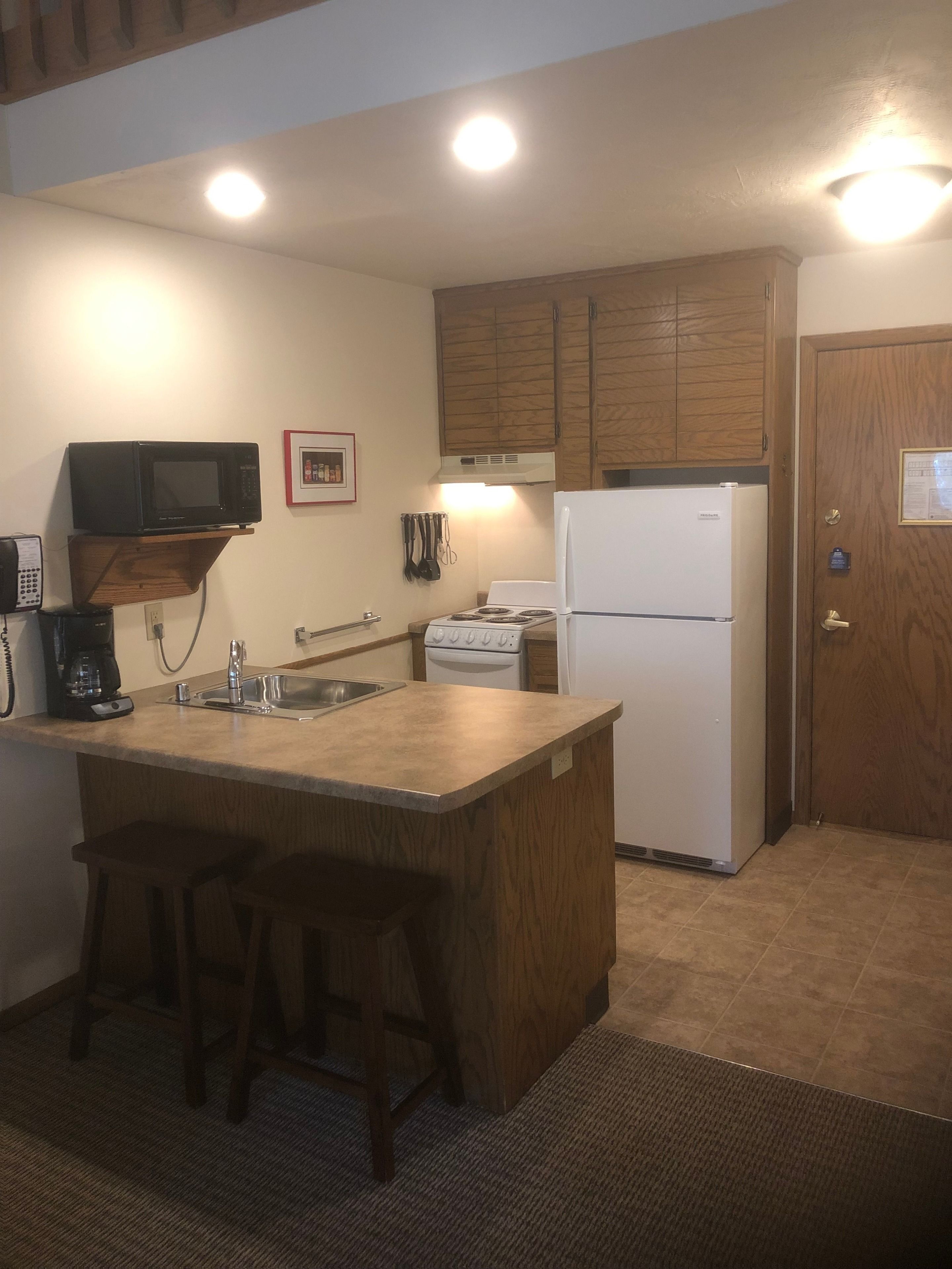 A compact kitchen with a built-in microwave, a stove, and a refrigerator. There are wooden cabinets and a small bar stool.
