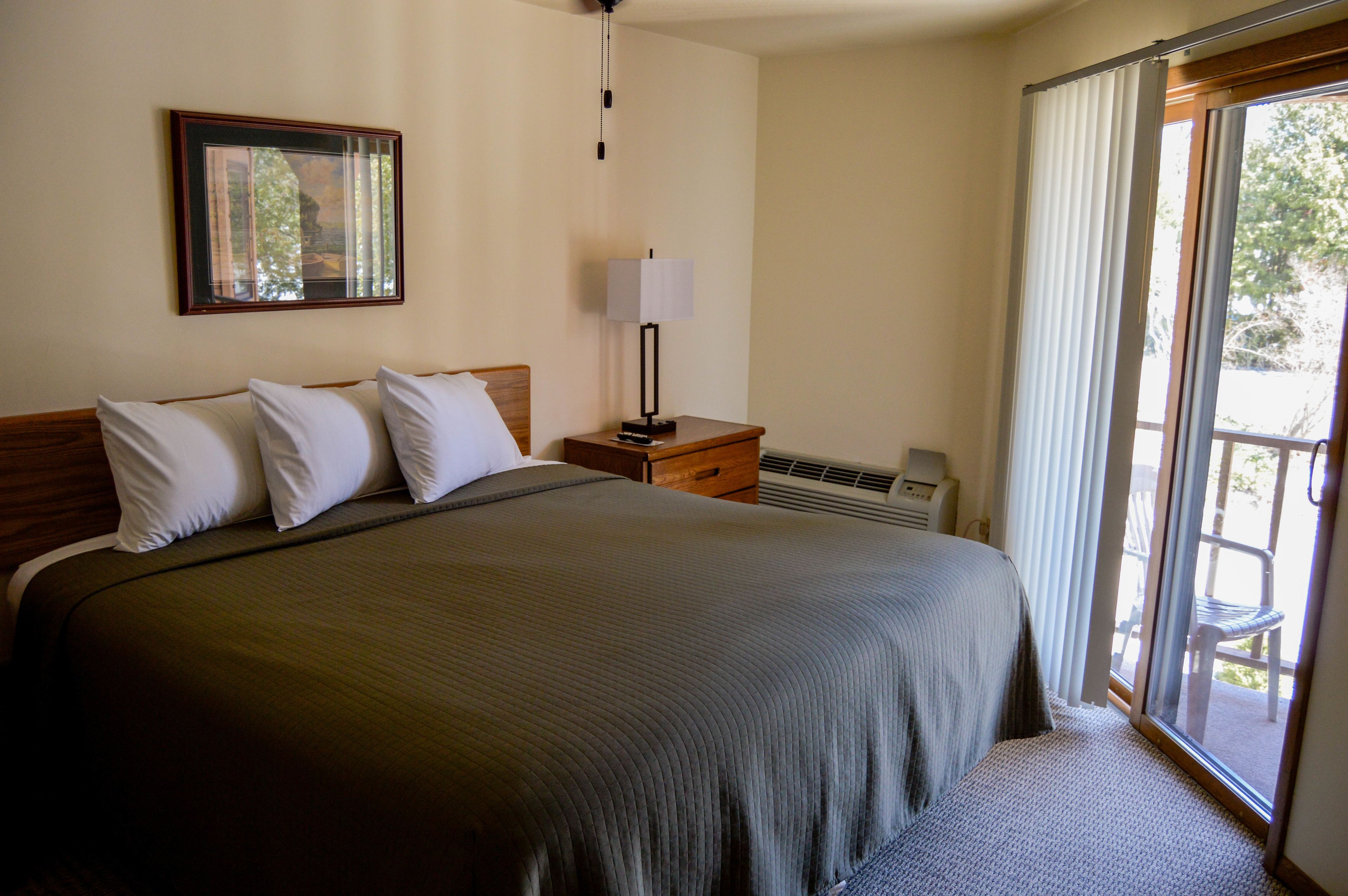 A hotel room with a large bed, a nightstand, a lamp, and a sliding glass door to the outside.