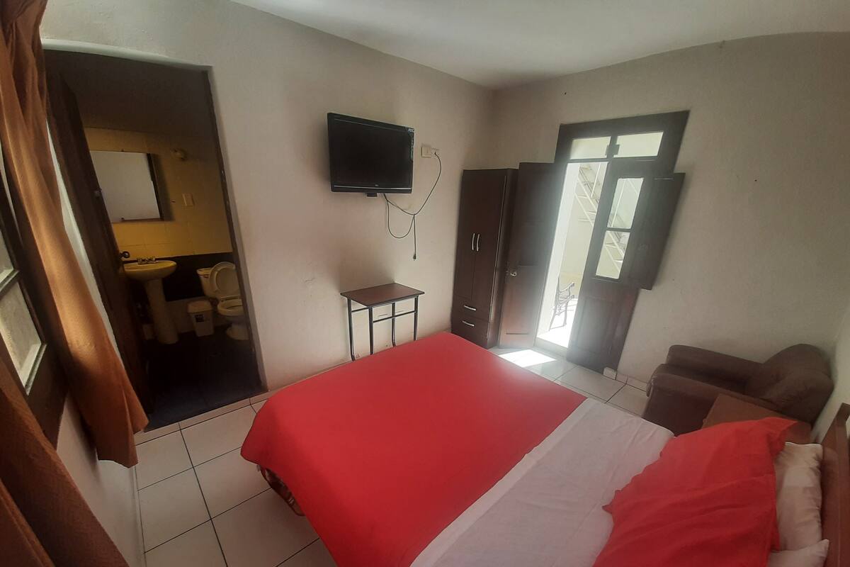 Un dormitorio con cama, televisor montado en la pared, armario y baño visible a través de una puerta abierta.