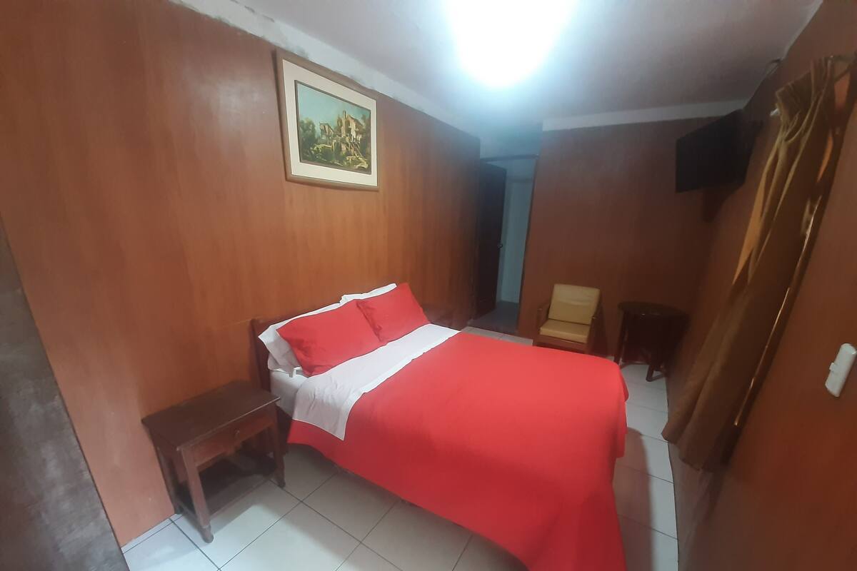 Un dormitorio con una cama, un banco de madera, una silla, un televisor y un cuadro en la pared.