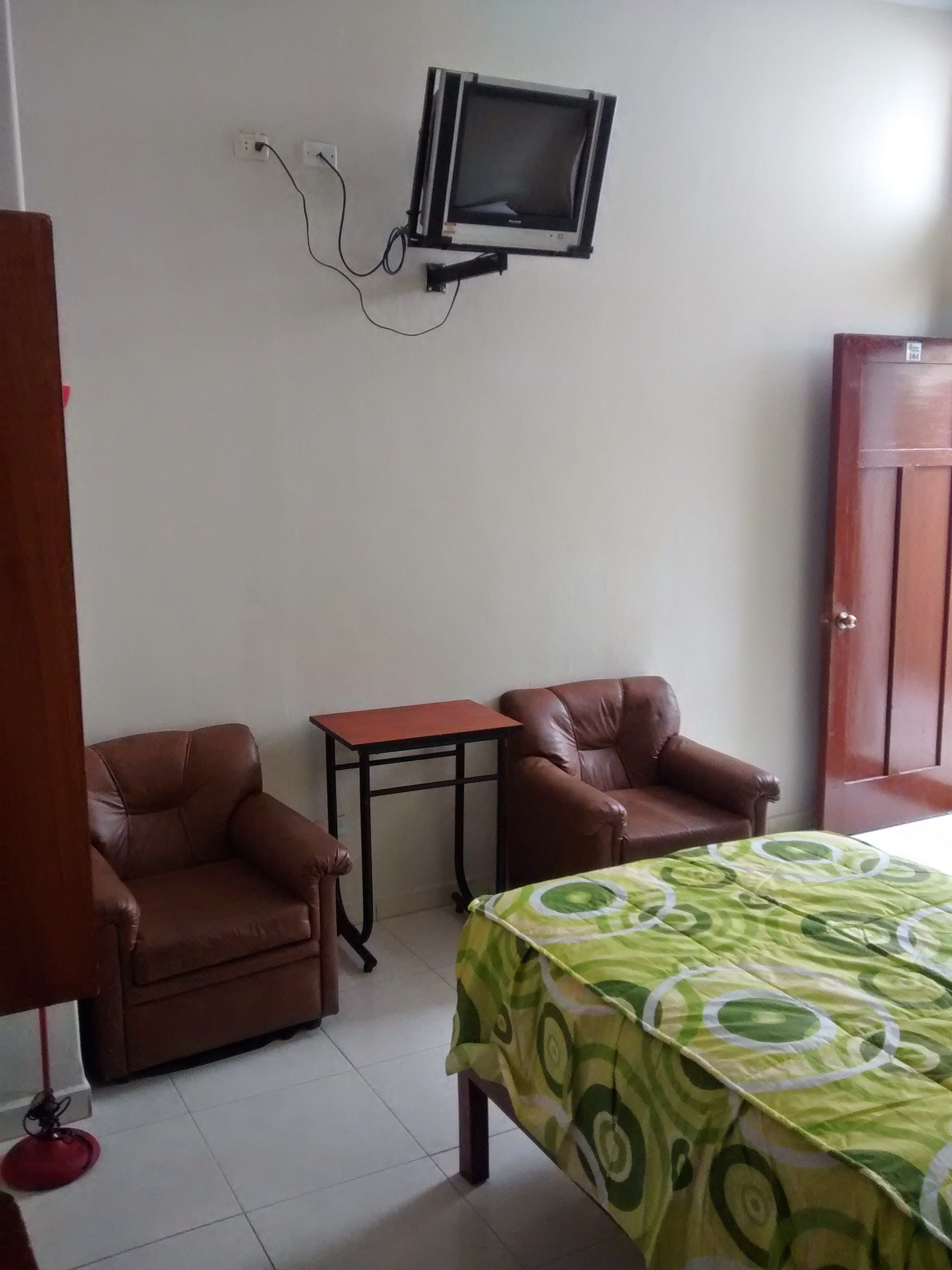 A room with two brown leather chairs, a small table, a bed with a green patterned bedspread, and a mounted television on the wall.