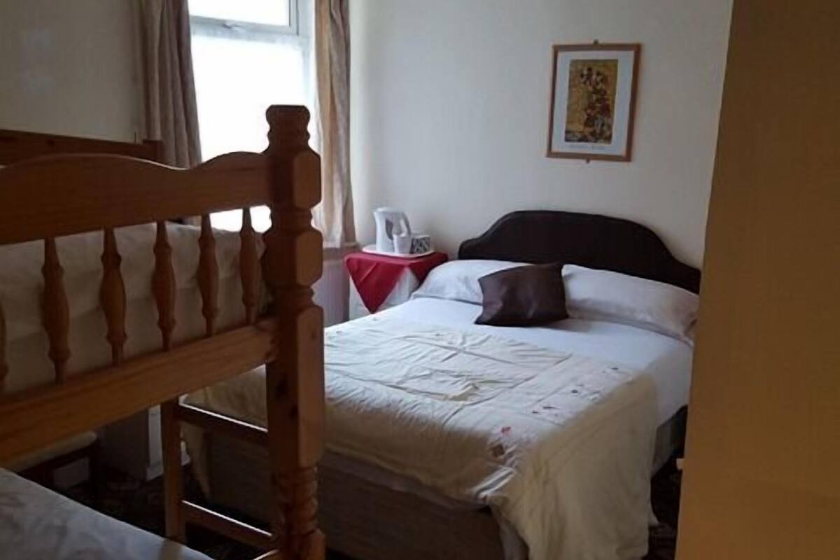 A bedroom with a wooden headboard, a bed with white bedding, a red table with a white object on it, and a framed picture on the wall.