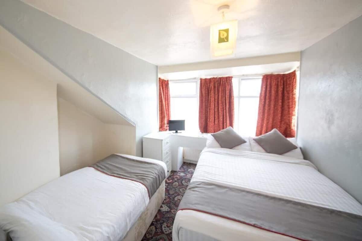 A small, well-lit room with two single beds, a desk with a computer, and a window with curtains.